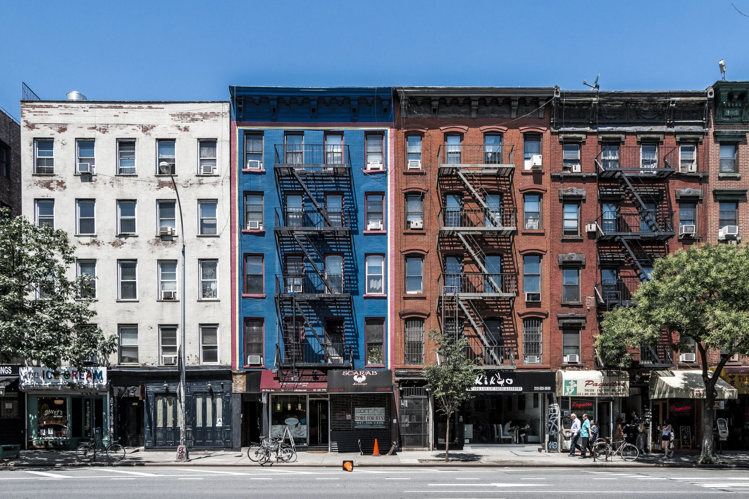 Colorful city buildings with fire escapes, storefronts, bicycles, and pedestrians on the sidewalk under a clear blue sky.