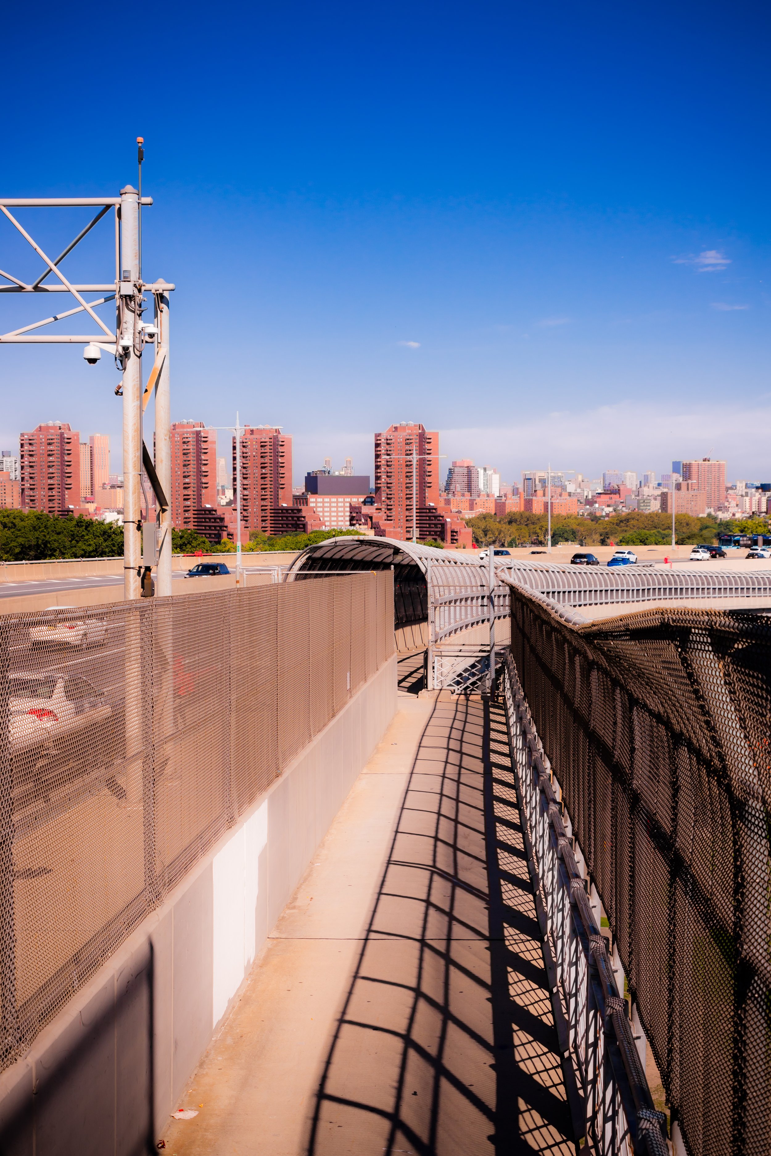 View of a city skyline with tall red-brick buildings, a pedestrian bridge with shadows, and a clear blue sky.