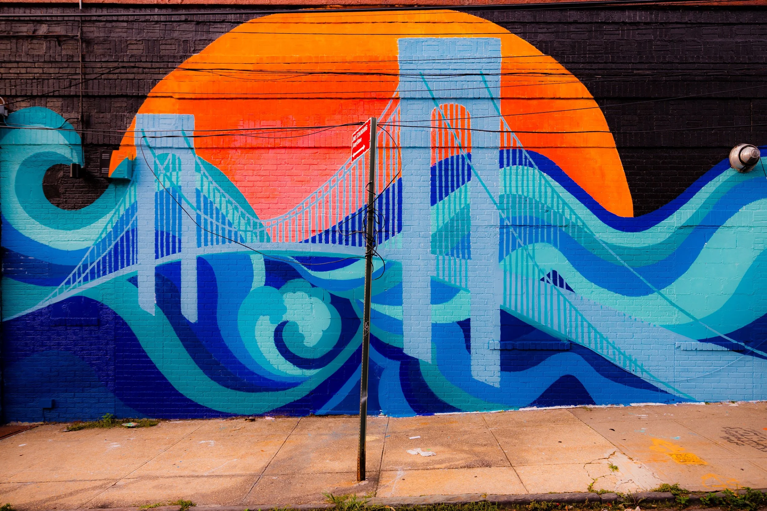 Colorful mural on brick wall depicting a bridge over stylized waves with a large orange circle background.