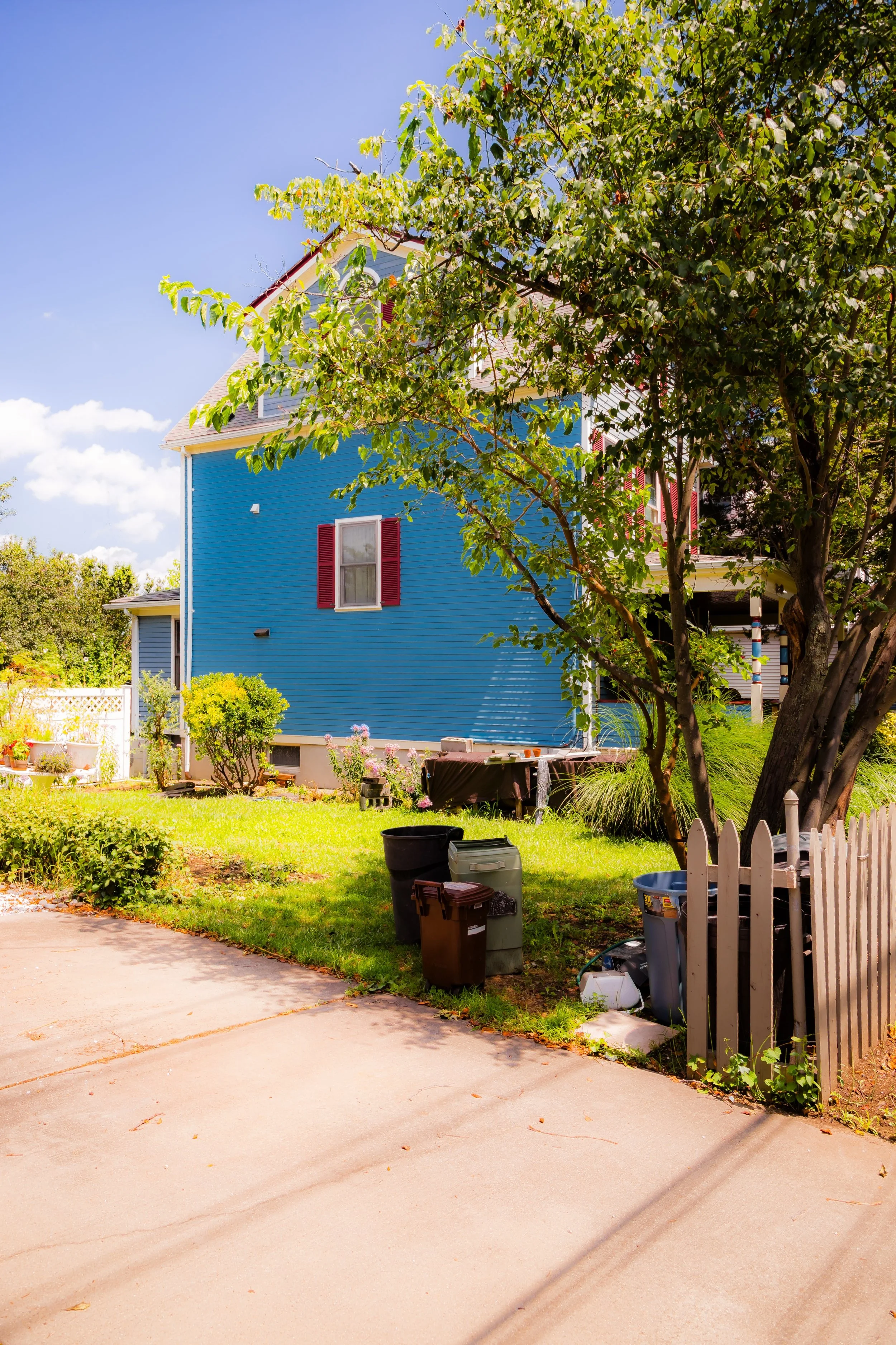 A blue house with red shutters and a tree in front, surrounded by a green yard with potted plants and trash bins, under a partly cloudy sky.