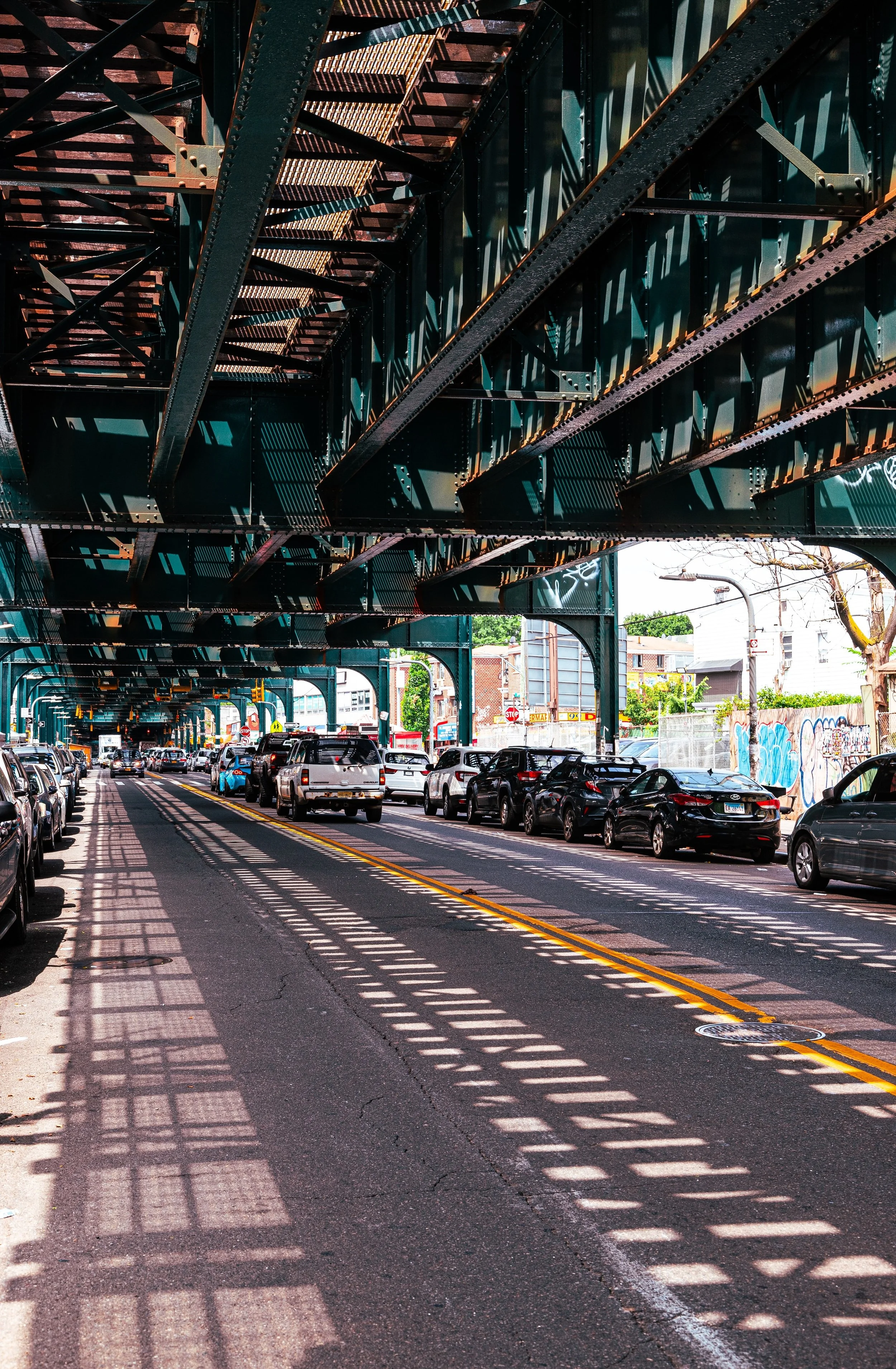 View of a street under an elevated train track, with parked cars on both sides and shadows cast by the track's structure on the road.