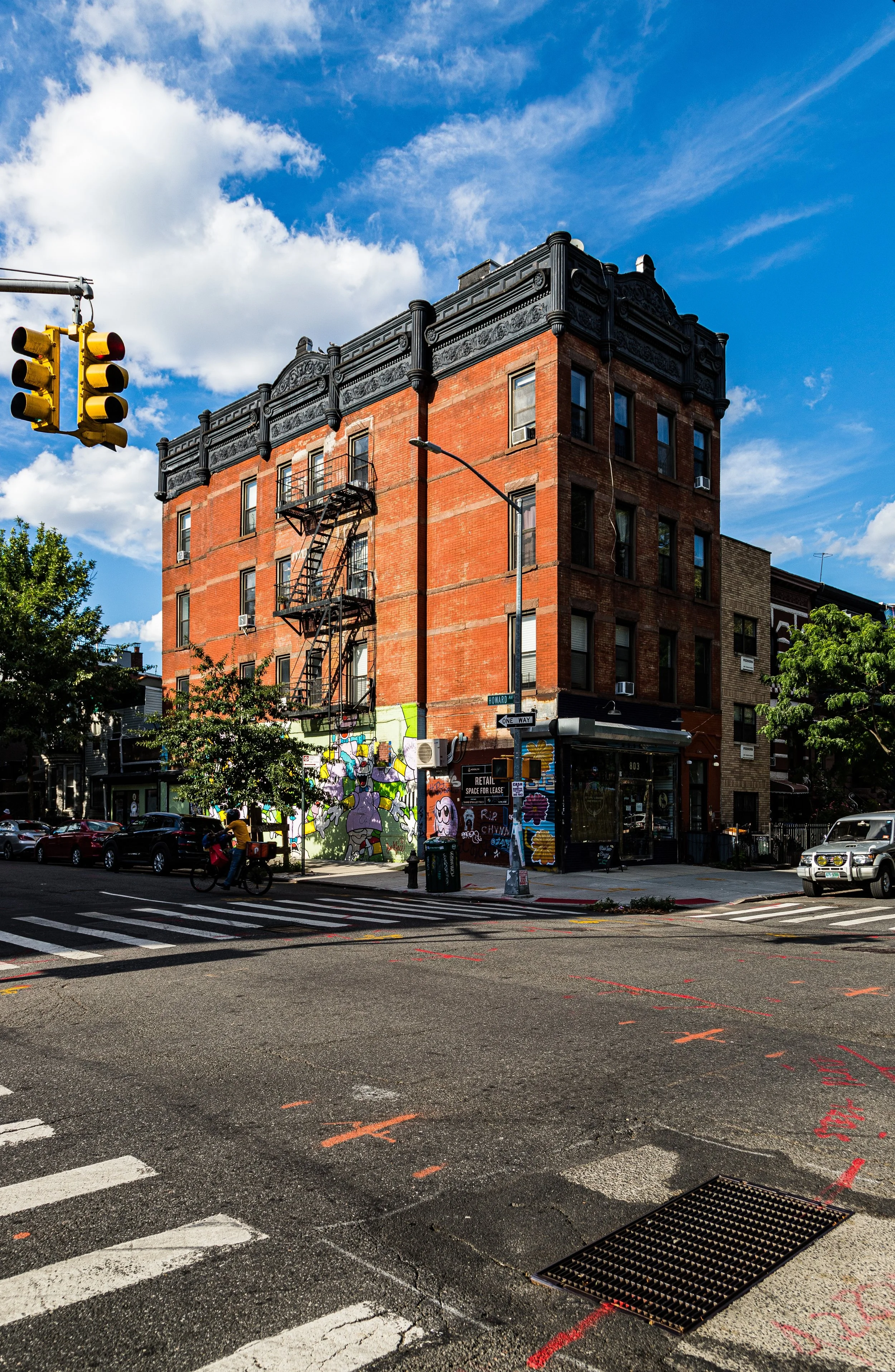 A four-story red brick building on a city street corner with a fire escape, graffiti art, and a retail space for lease, under a bright blue sky with clouds.
