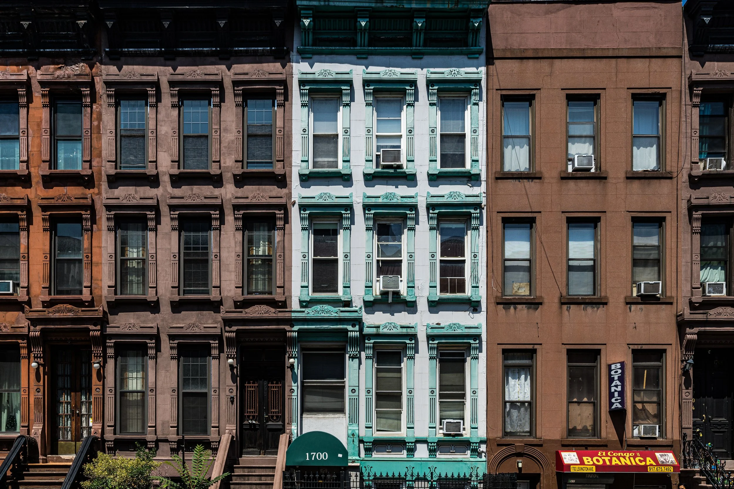 Row of three colorful urban apartment buildings, with brown, teal, and reddish-brown facades, featuring decorative window frames, some air conditioning units, and storefronts at street level.