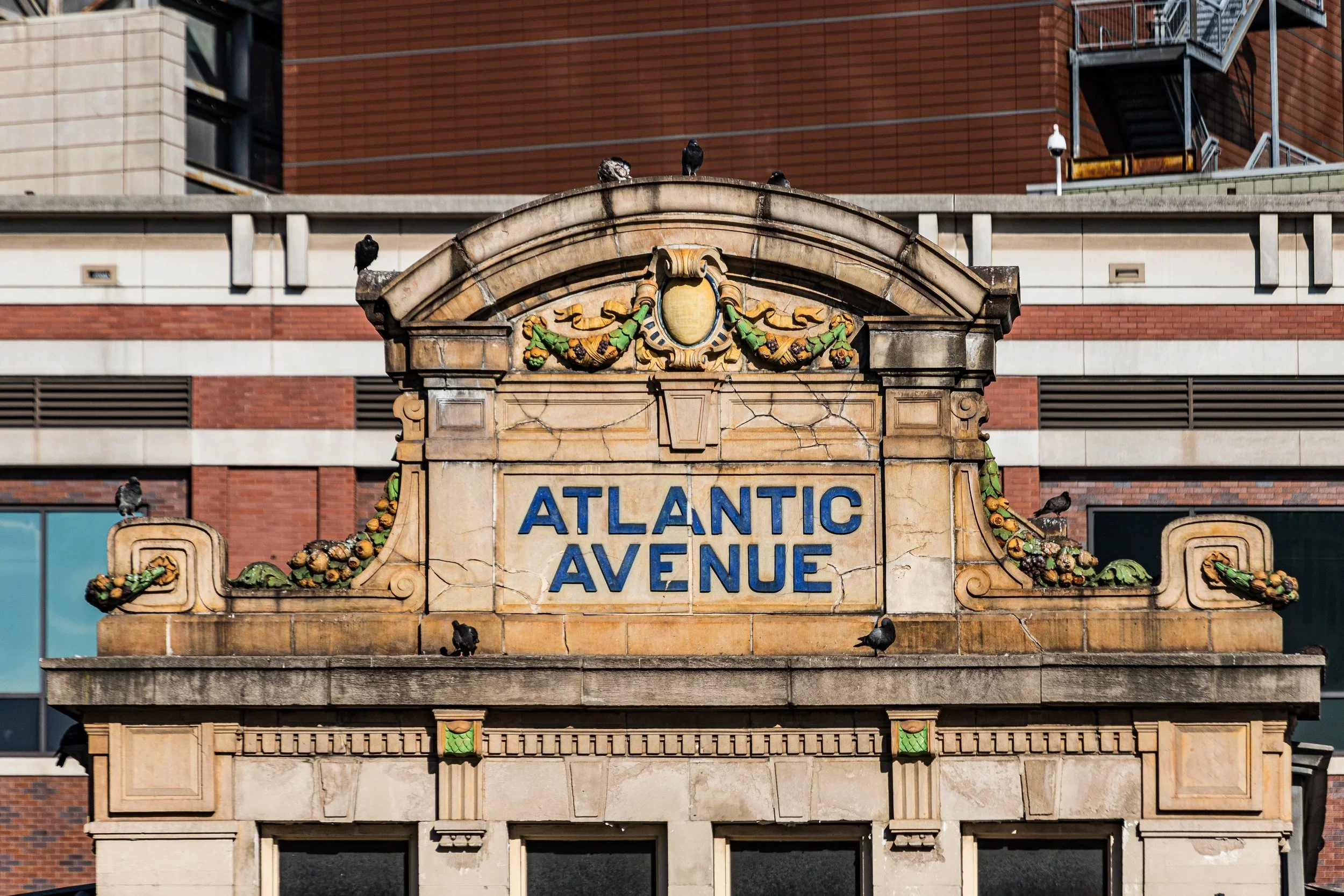 Sign reading 'Atlantic Avenue' with decorative floral and garland accents, pigeons perched on top and around the sign.