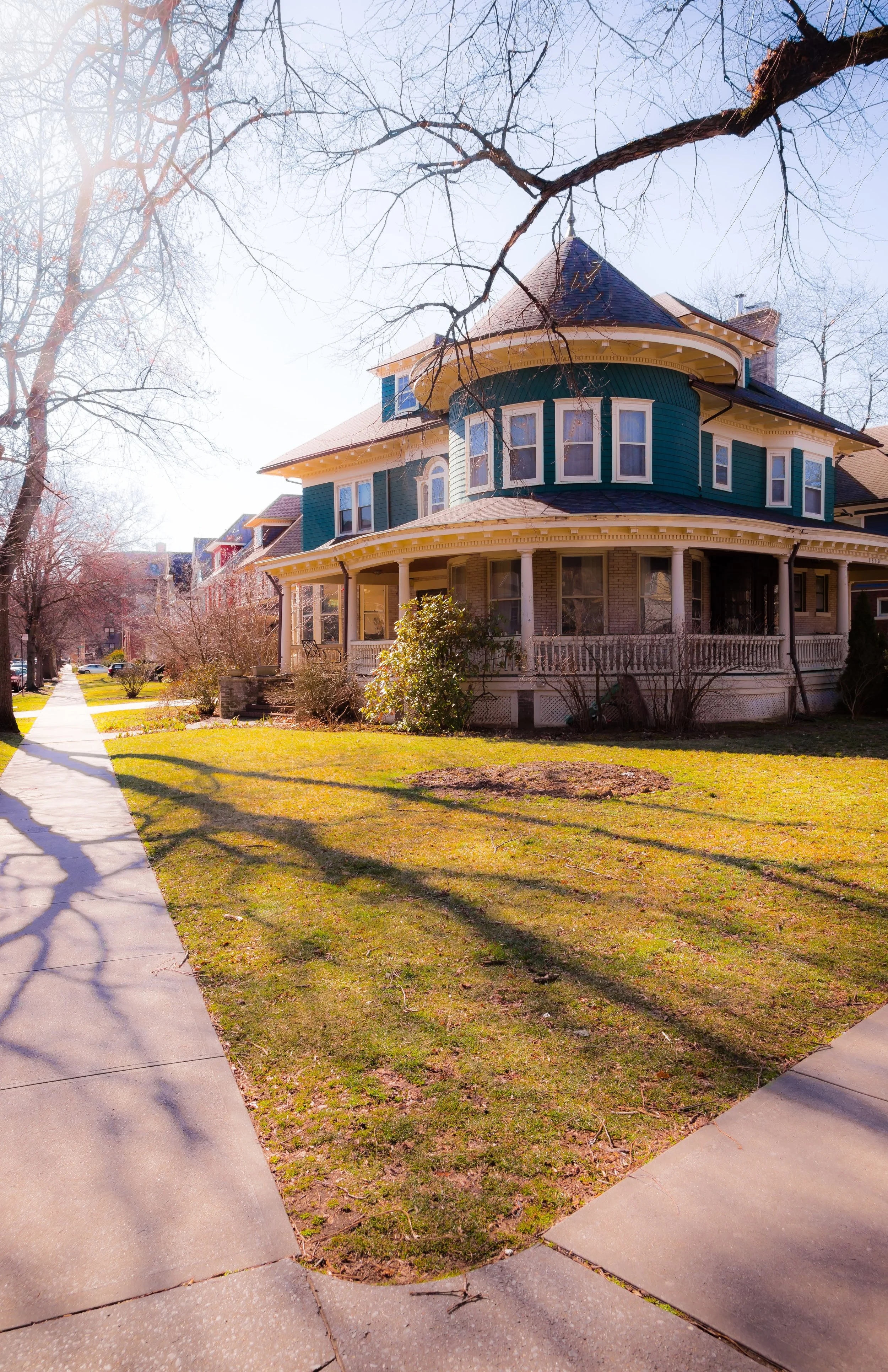 A large Victorian-style house with a rounded turret, teal siding, and a wrap-around porch, situated on a grassy lawn with trees and a sidewalk in a residential neighborhood on a sunny day.