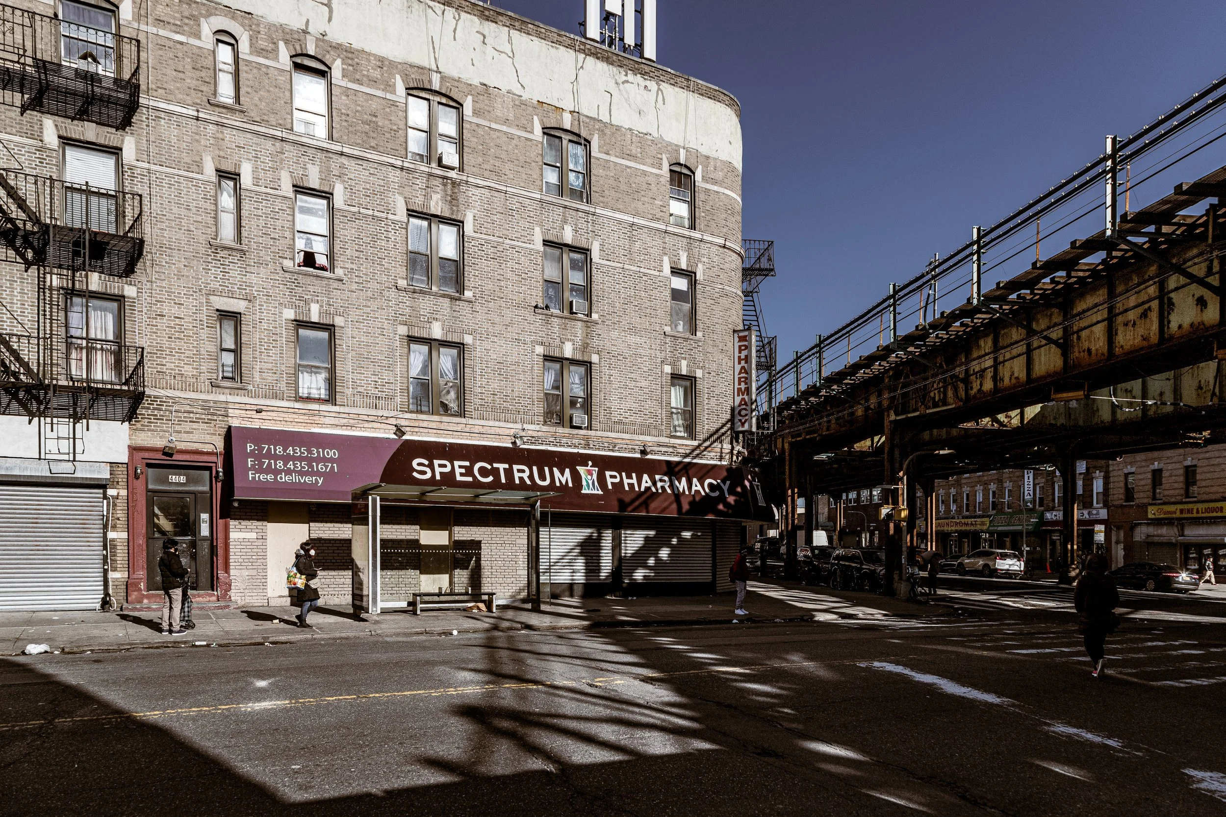 Street scene with a building and an elevated train track overhead. A pharmacy storefront labeled "Spectrum Pharmacy" with phone numbers and notice of free delivery. Pedestrians walk on the sidewalk, some wearing masks, and cars are parked along the street. Shadows cast across the road from the elevated tracks.