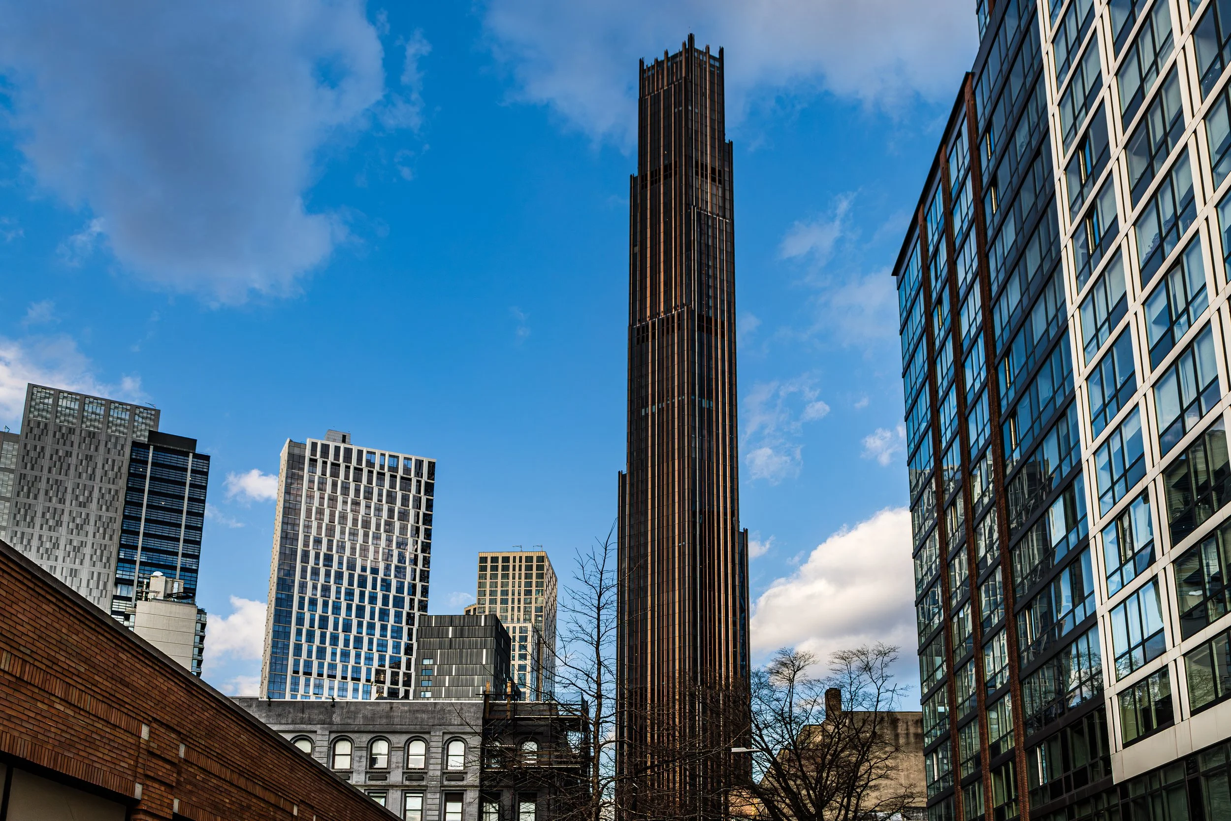 City skyscrapers with a prominent tall dark building in the center against a partly cloudy blue sky.