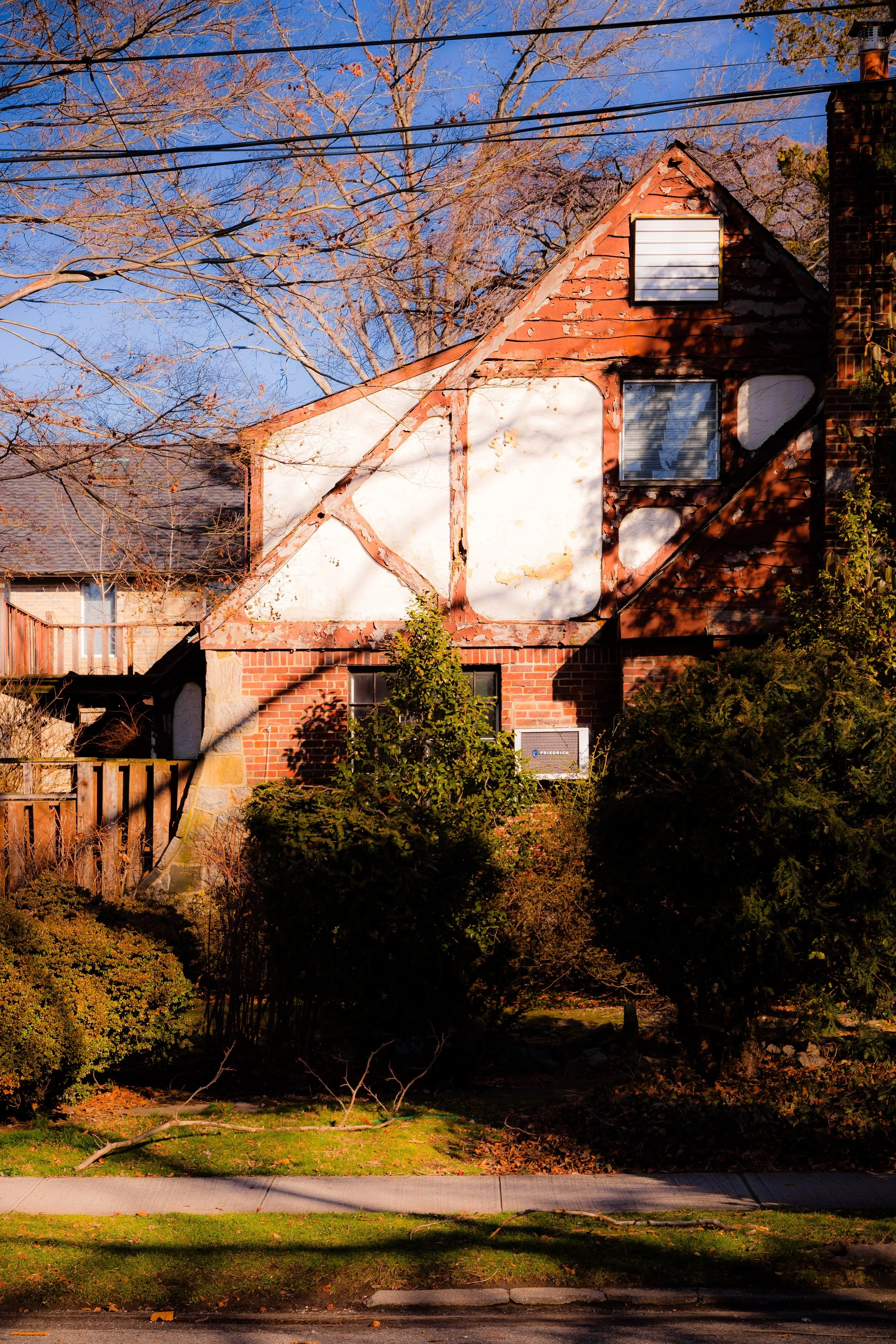 An old brick house with a steep triangular roof, showing signs of wear with peeling paint. There are two windows visible, one small vent at the top and one large window below. The house is partially obscured by trees and bushes, with a sidewalk in front and power lines overhead.