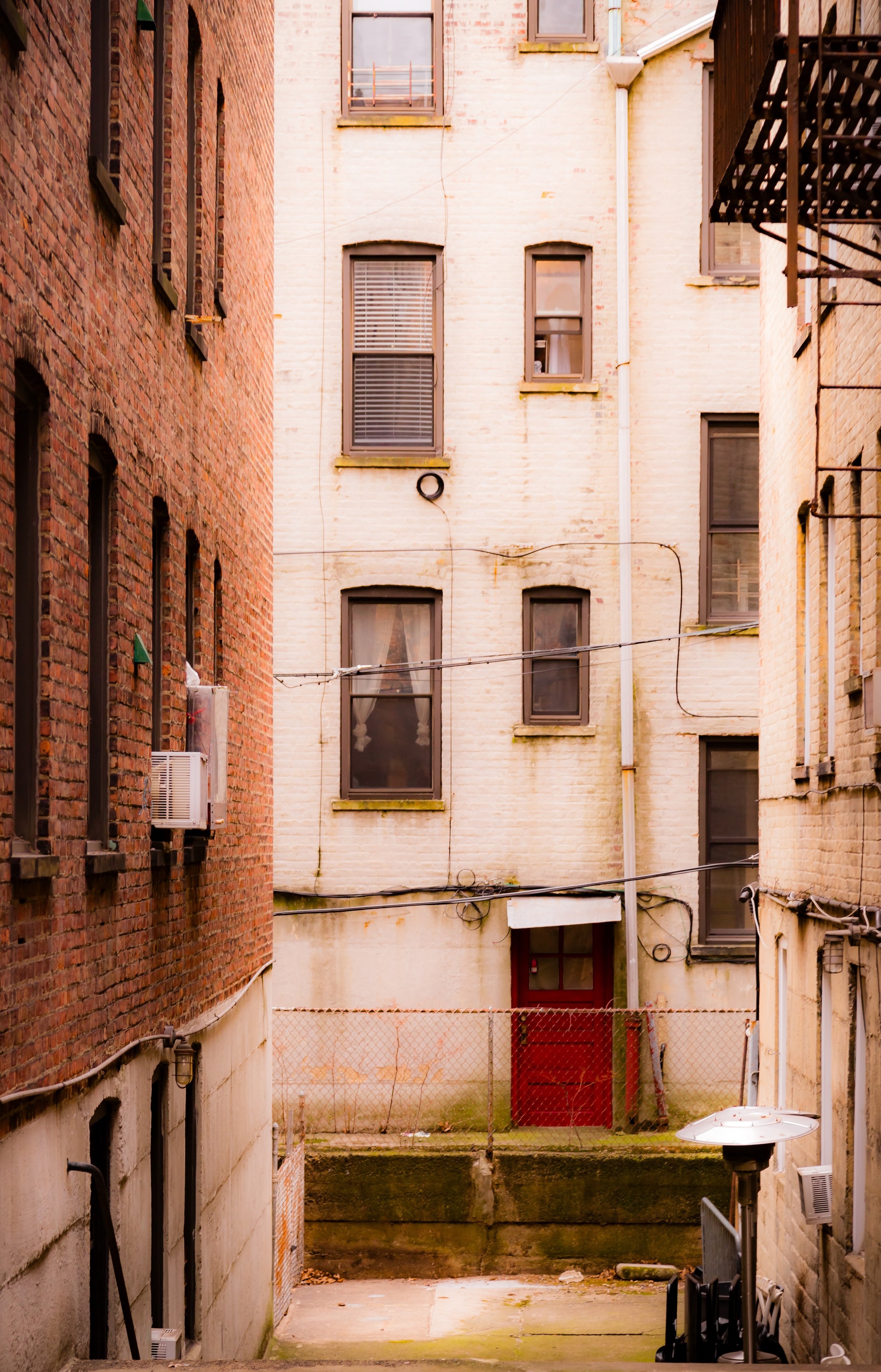View of a narrow alley with brick apartment buildings on each side, multiple windows, and an enclosed backyard with a fenced area and outdoor equipment.