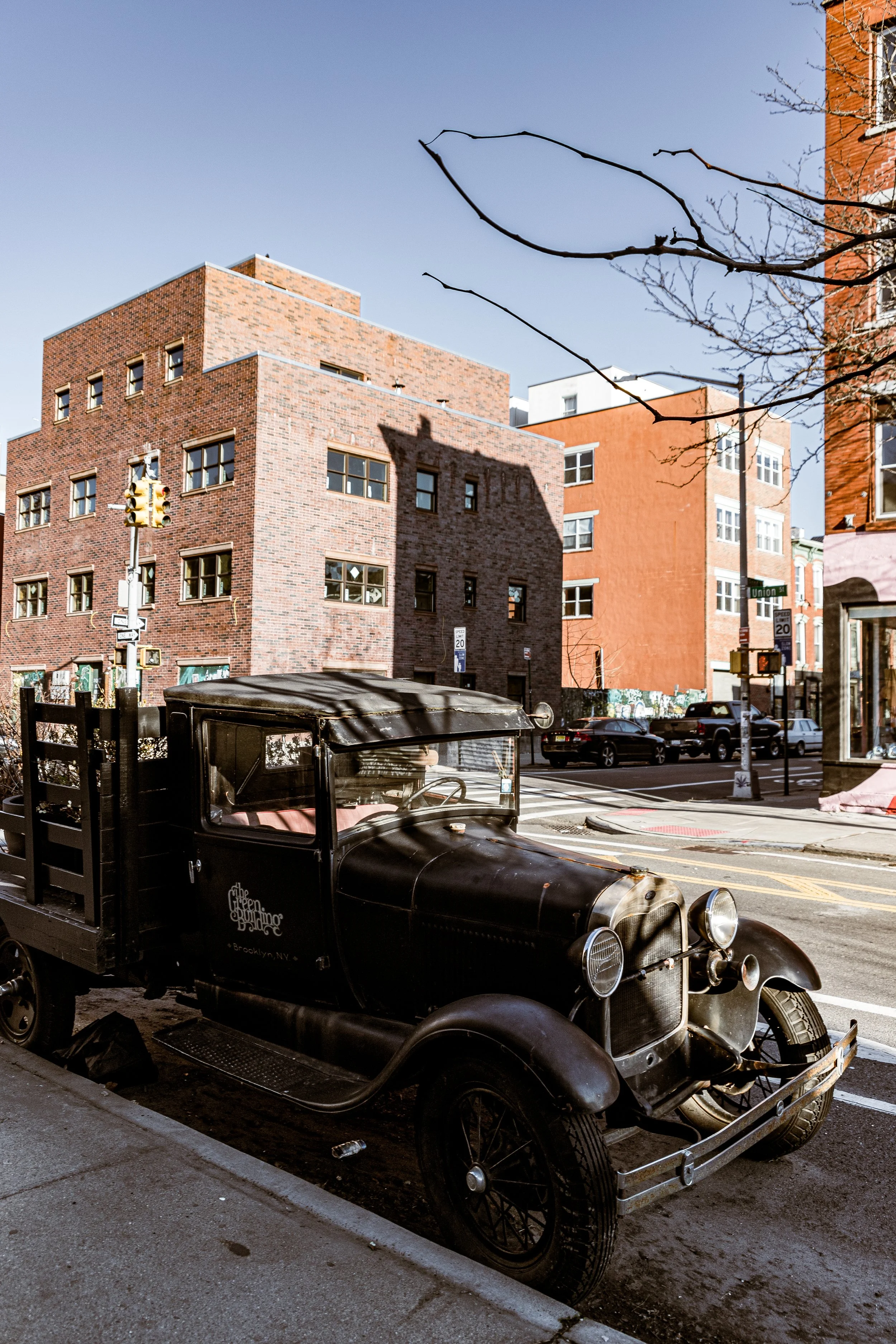 An old black vintage truck parked on a city street with brick buildings in the background.