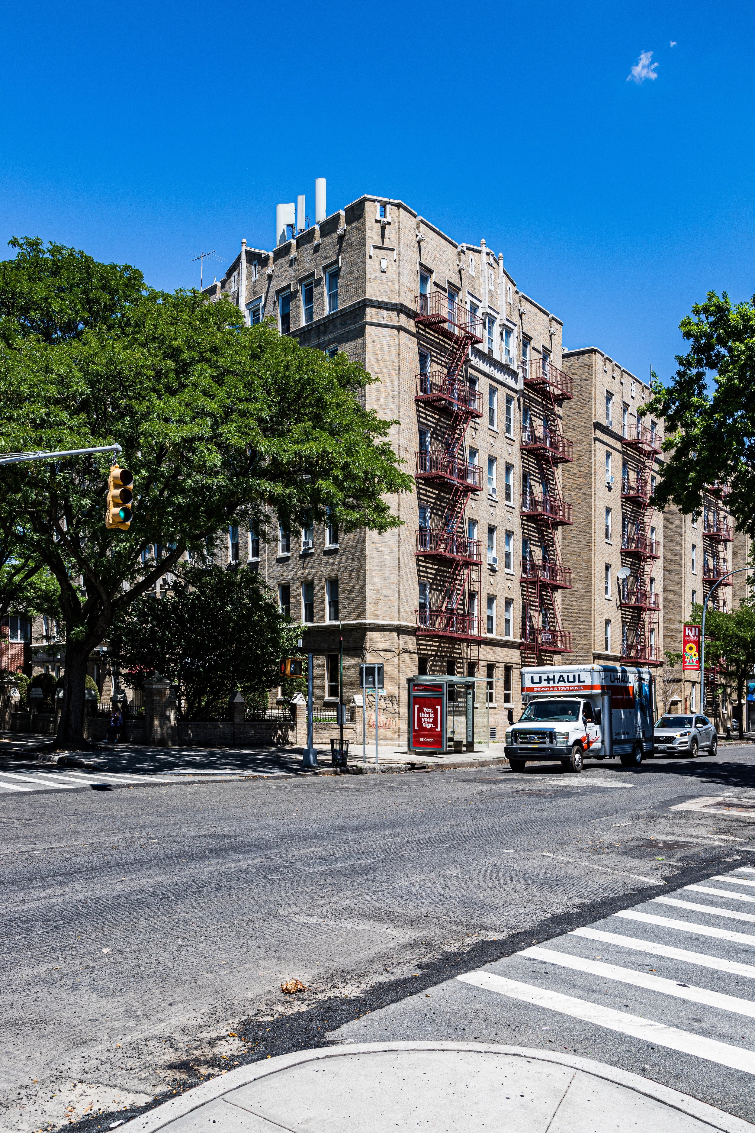A city street corner with a large brick apartment building, green trees, cars, a U-Haul truck, and a traffic light against a clear blue sky.