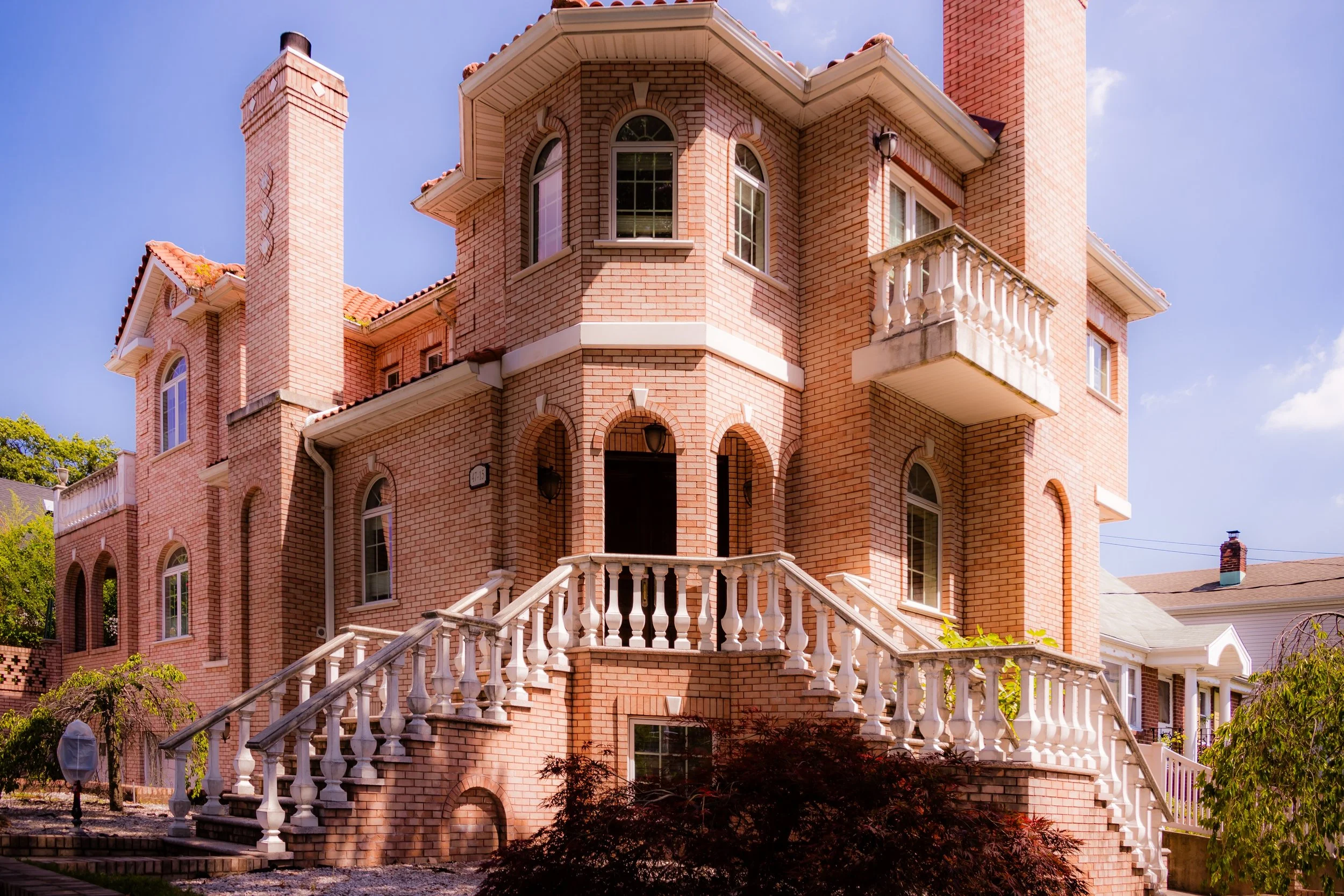 A large brick house with multiple stories, arched windows, a chimney, and a decorative balcony. Stone stairs lead up to the front entrance.