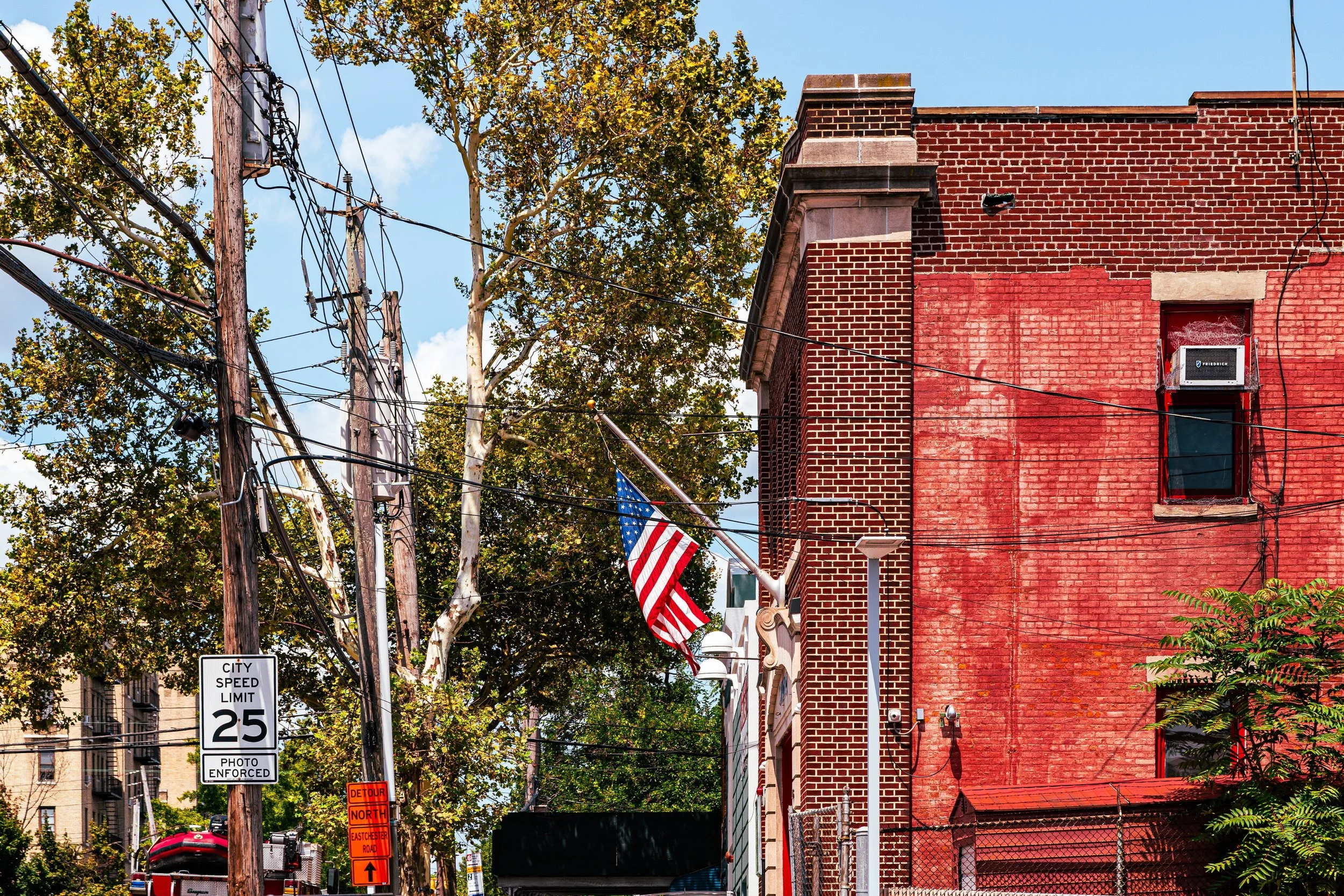 This is a city street scene featuring a red brick building with a window air conditioning unit, power lines, and a flagpole with an American flag. There are trees with green leaves, a blue sky, and street signs indicating a 25 mph speed limit and a detour.