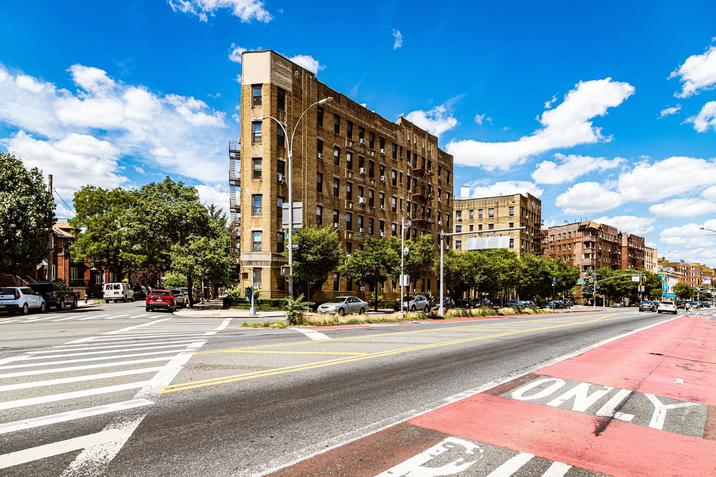 City street with multi-story brick buildings, trees, parked cars, crosswalks, and a blue sky with scattered clouds.