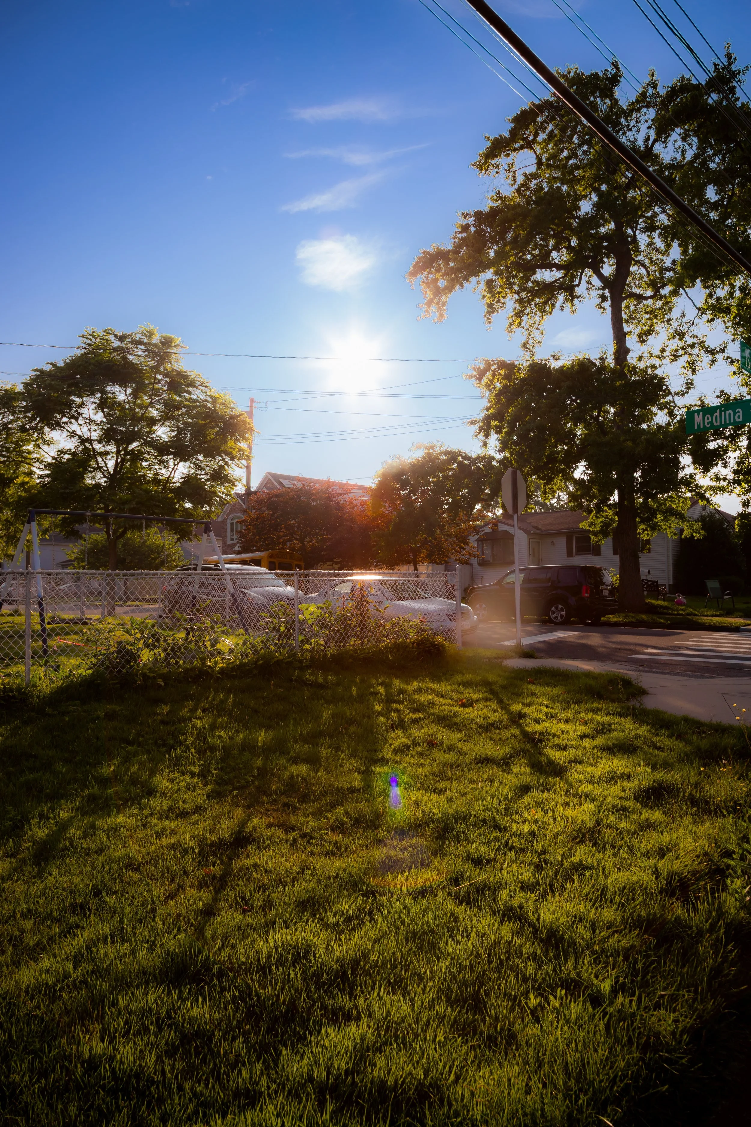 Sun shining over a suburban neighborhood with trees, houses, parked cars, a chain-link fence, and a street sign at the intersection of Medina Street during late afternoon.