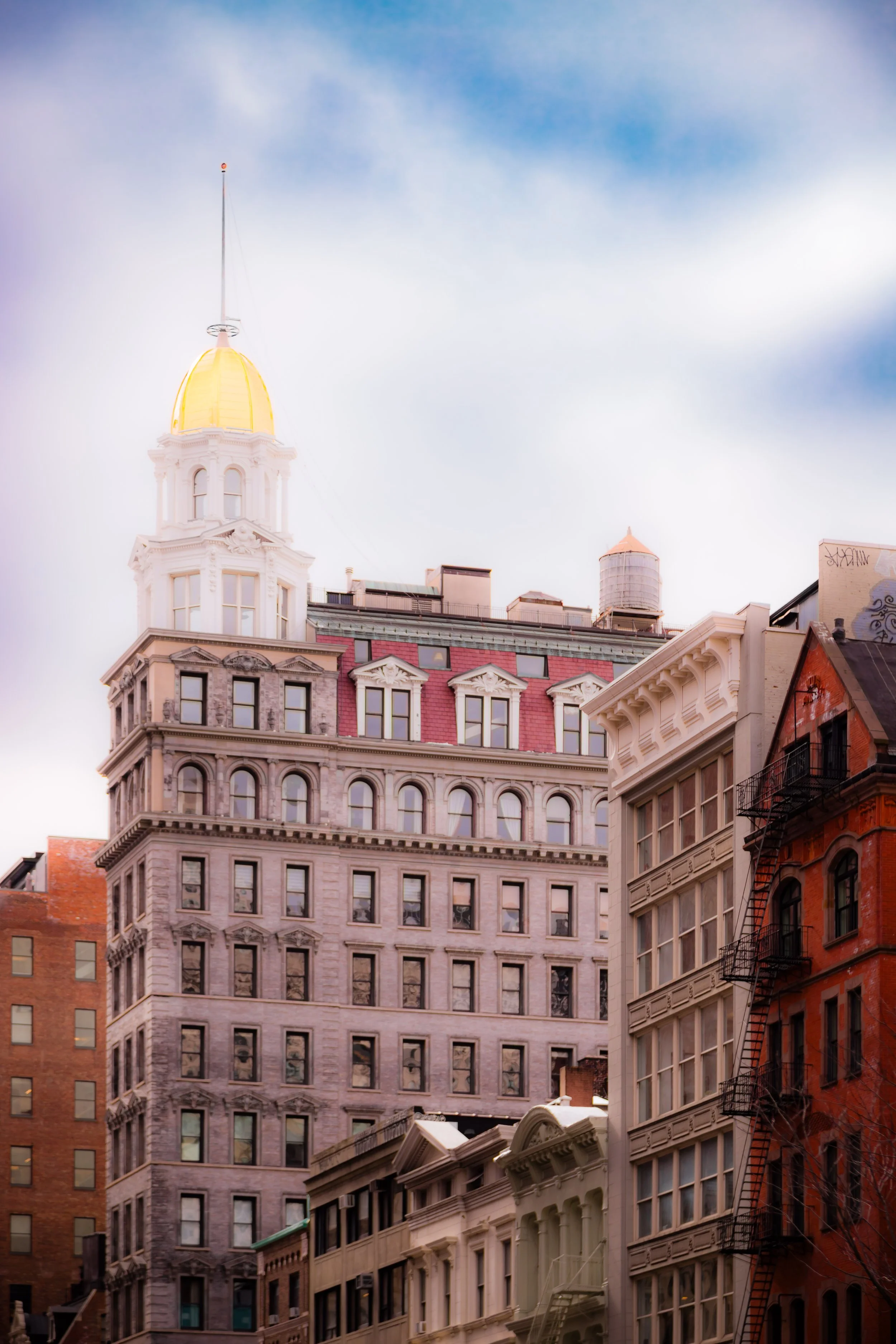 Cityscape with historic multi-story buildings with ornate facades, fire escapes, and a golden-domed tower against a cloudy sky.