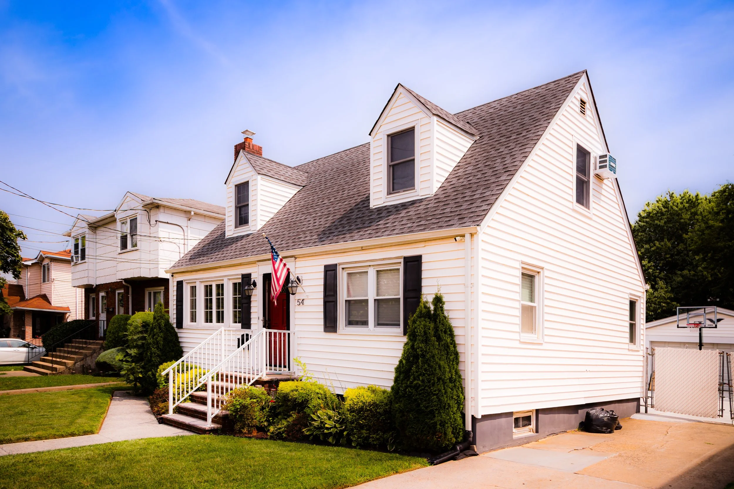 White two-story house with black shutters, front steps, American flag, and a small mountain of snow on the roof, in a neighborhood with other houses and a basketball hoop visible.