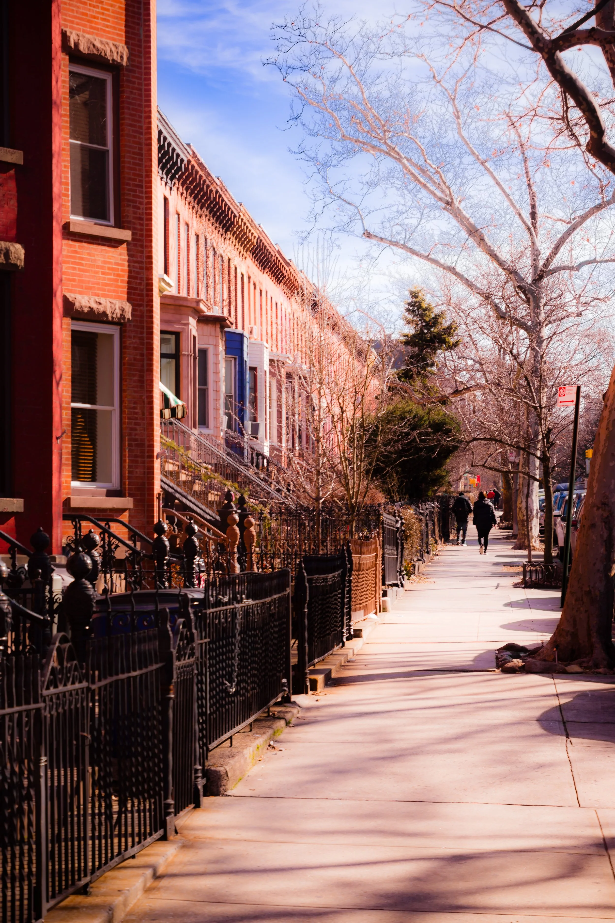 A sidewalk in an urban neighborhood with row houses on the left side, some with stairs and decorative railings, and leafless trees lining the sidewalk on the right. Two people are walking away in the distance on a sunny day with clear skies.