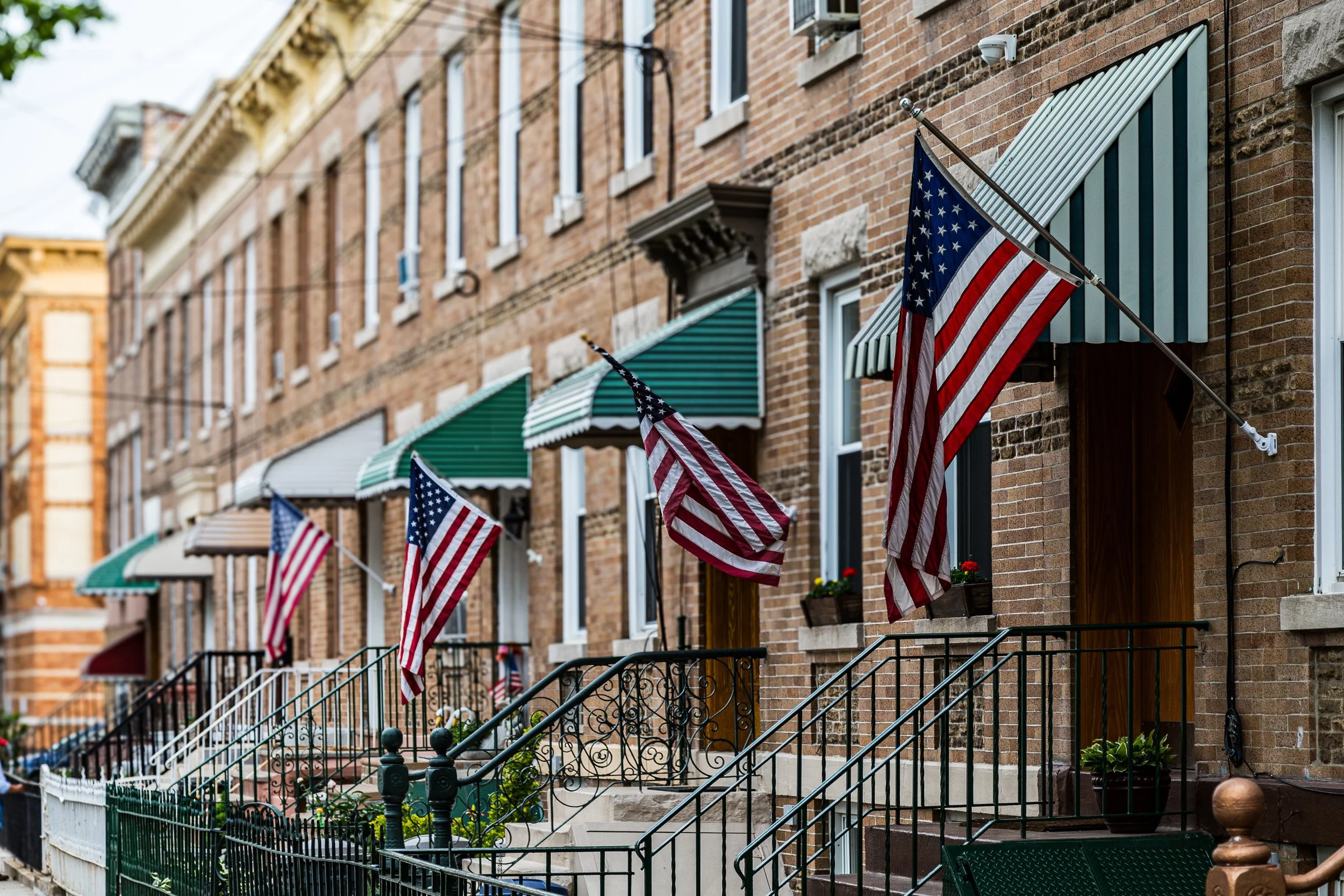 Row of brick townhouses with American flags hanging outside, green and striped awnings, and potted flowers on window sills.