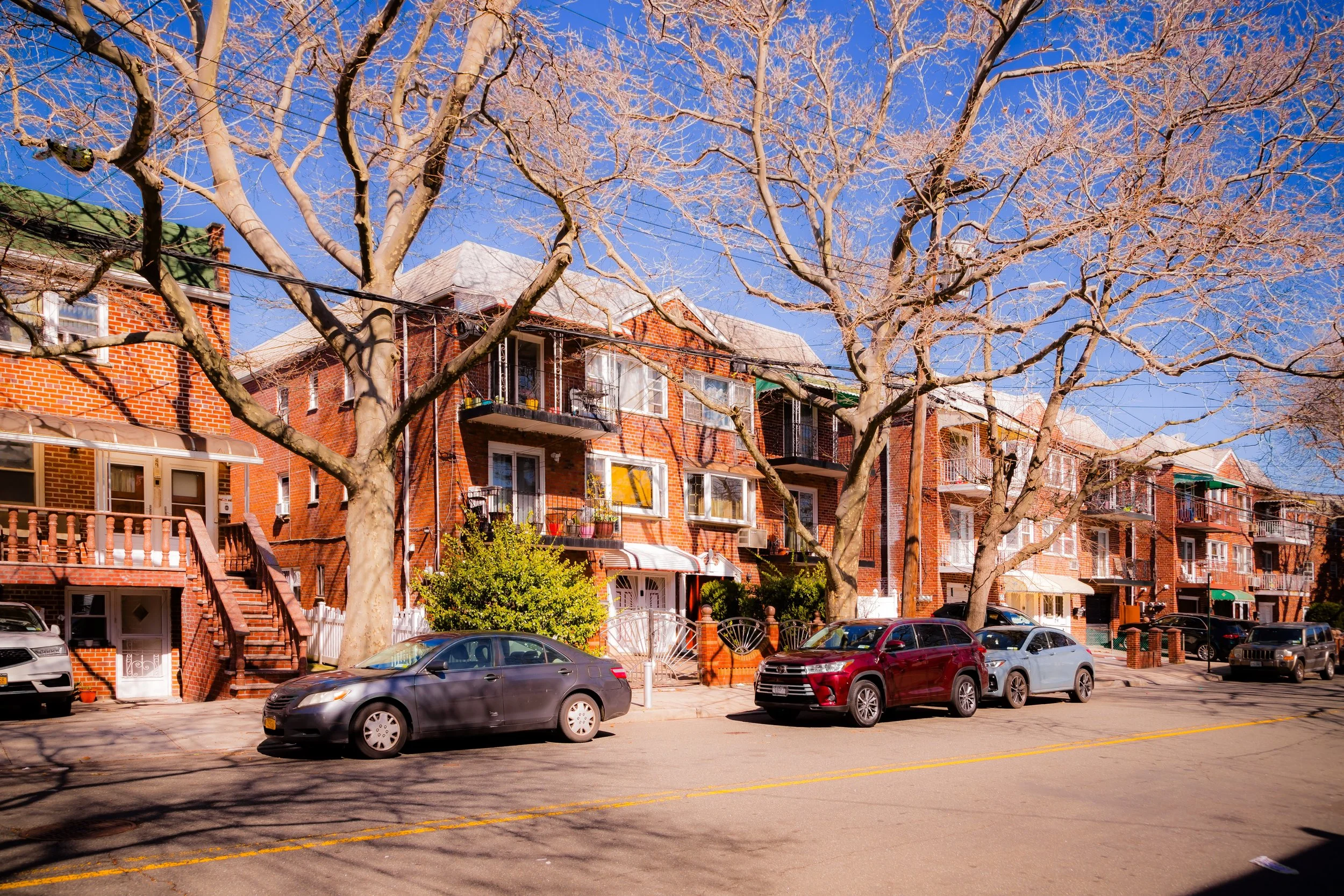 A street view with brick apartment buildings, leafless trees, parked cars, and a bright blue sky.