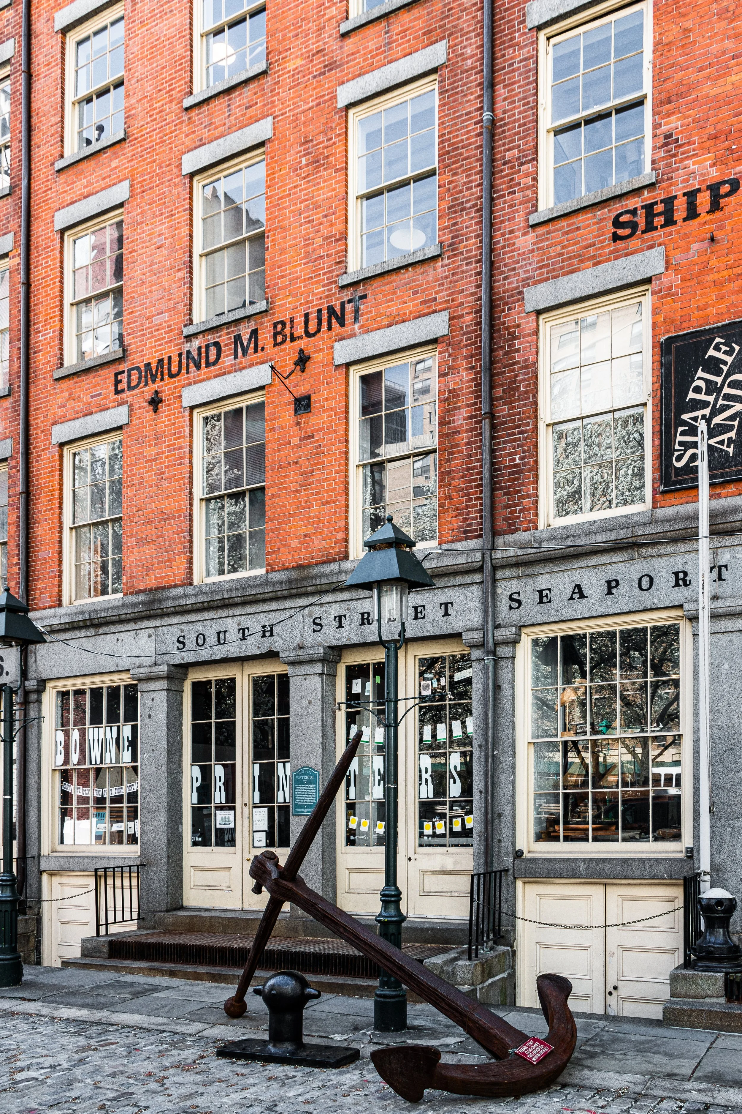 A brick building with multiple stories and large windows, marked with signs that read 'Edmund M. Blunt', 'South Street Seaport', and 'Ship'. In front of the building, there is a large decorative anchor leaning against a green street lamp post, and cobblestone pavement.