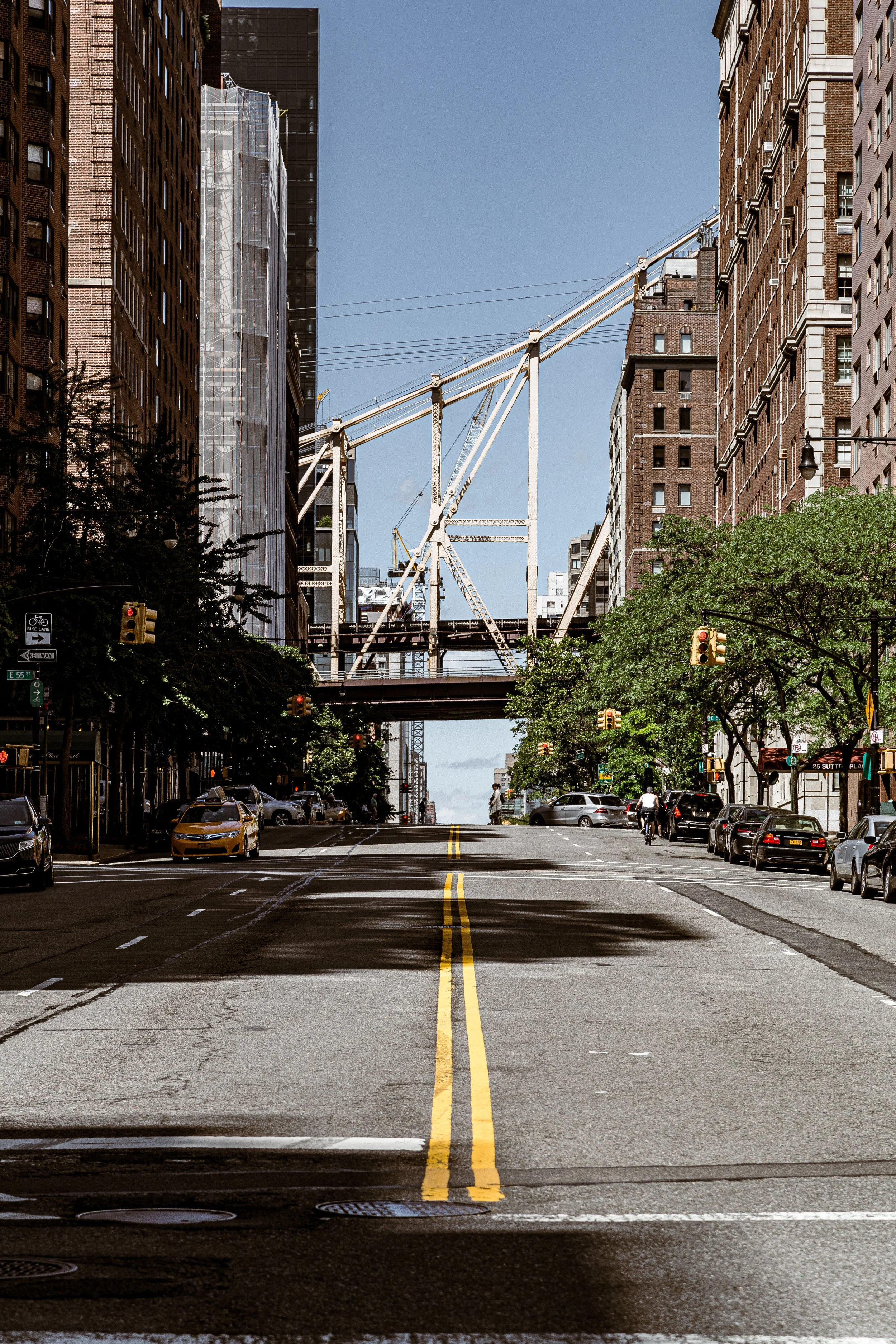 An empty city street with parked cars on both sides, tall buildings on either side, and a large construction crane overhead under a clear blue sky.