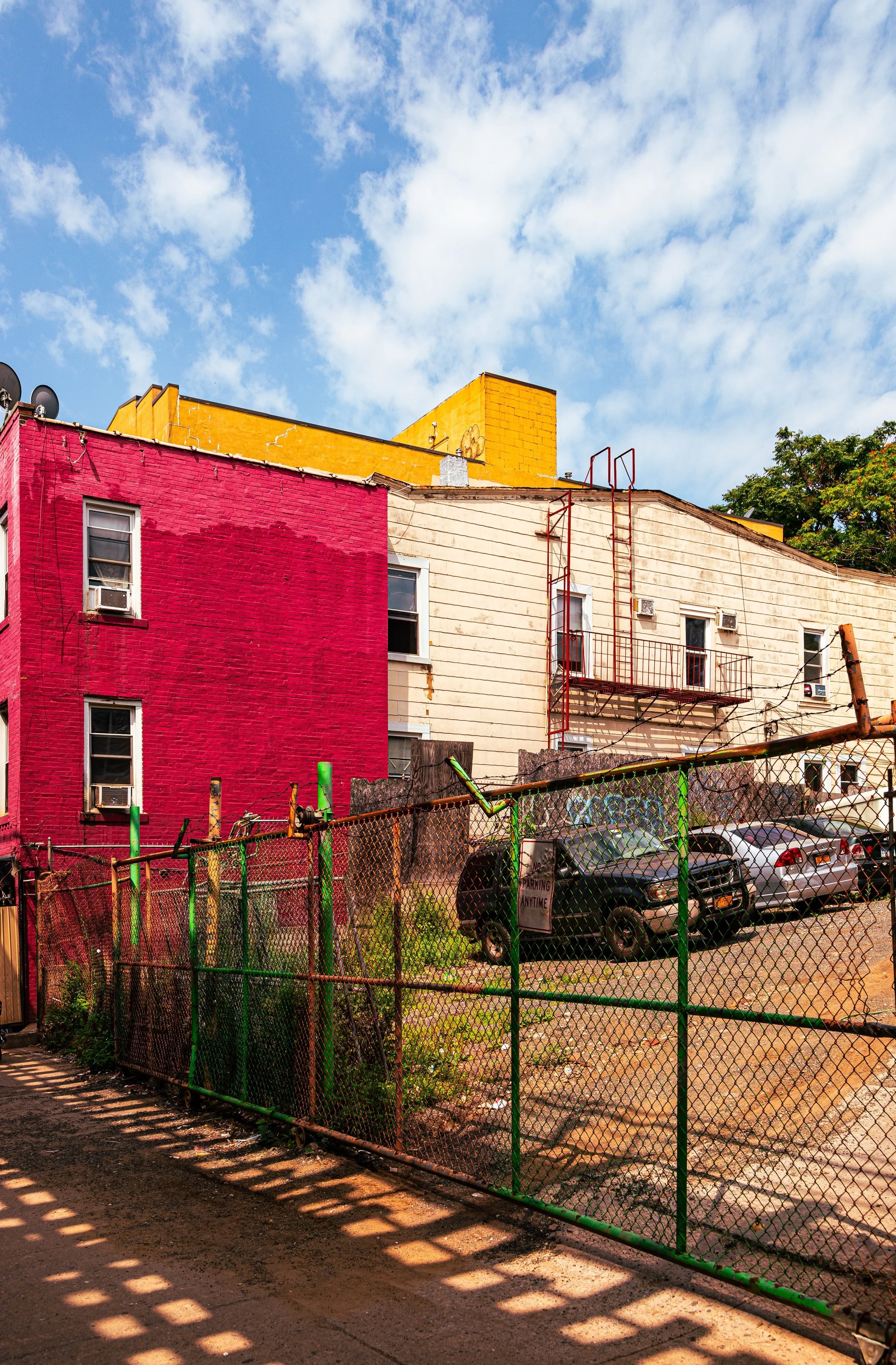 Colorful urban scene with a pink and white building, parking lot with cars, and a chain-link fence in the foreground, under a blue sky with clouds.