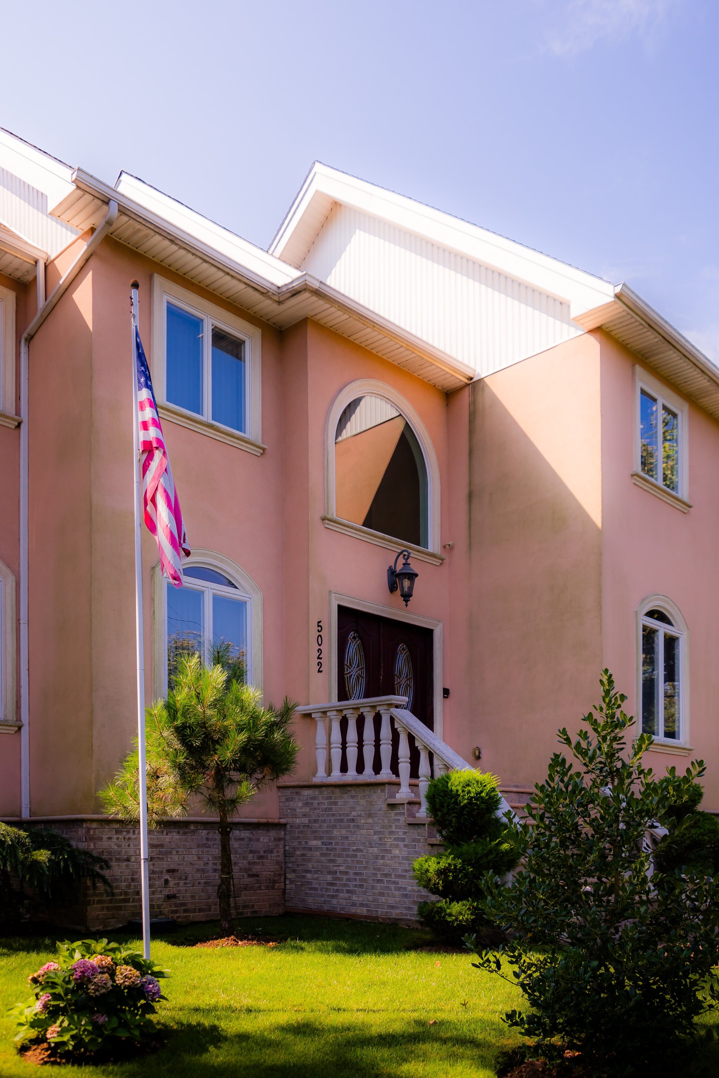 A pink multi-story house with white trim, a front porch with stairs, an American flag, landscaped yard with shrubs and flowers, and a clear blue sky.