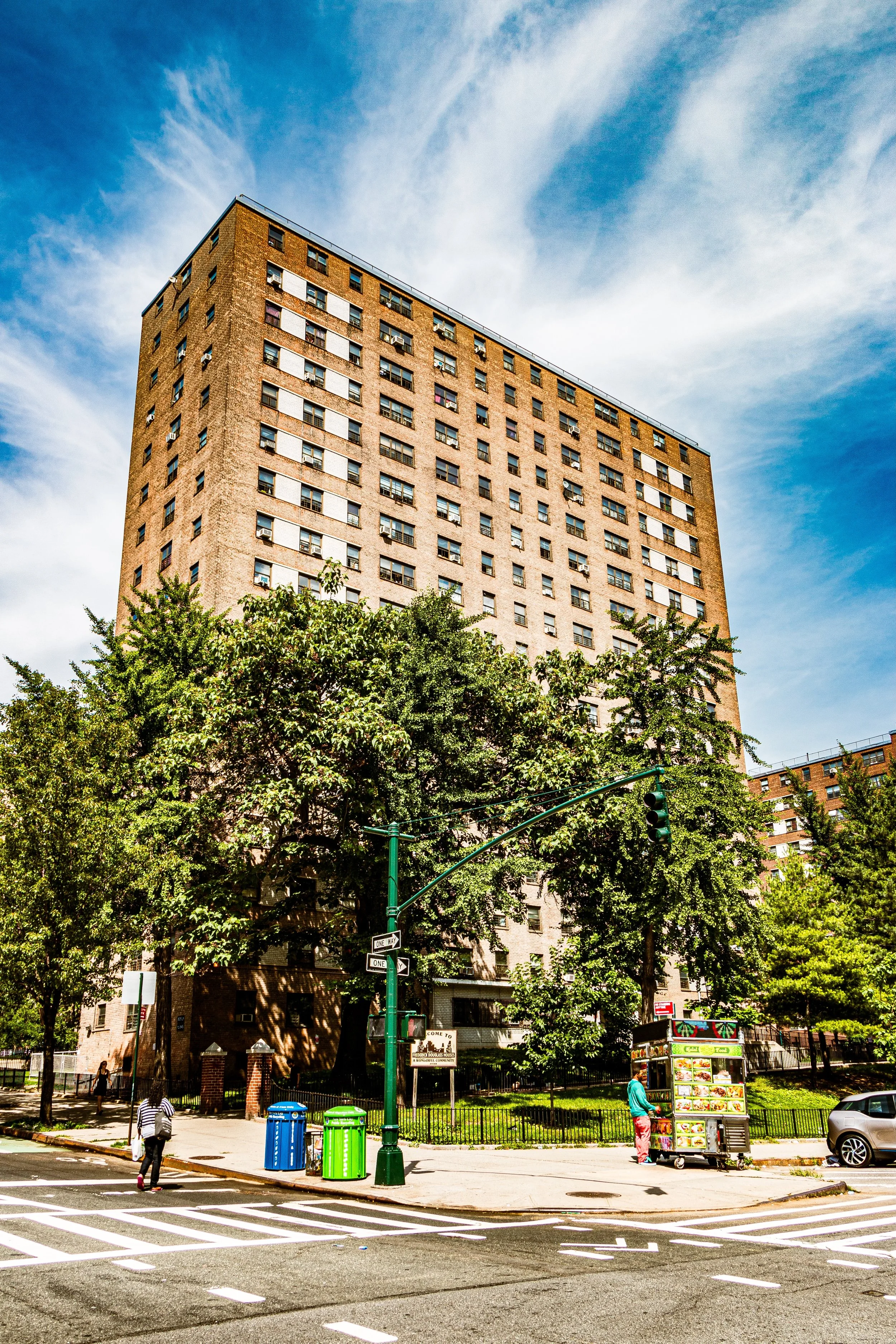 A tall brown apartment building with many windows, surrounded by trees, under a partly cloudy blue sky. In front, a street with a crosswalk, people, a food cart, and garbage bins.