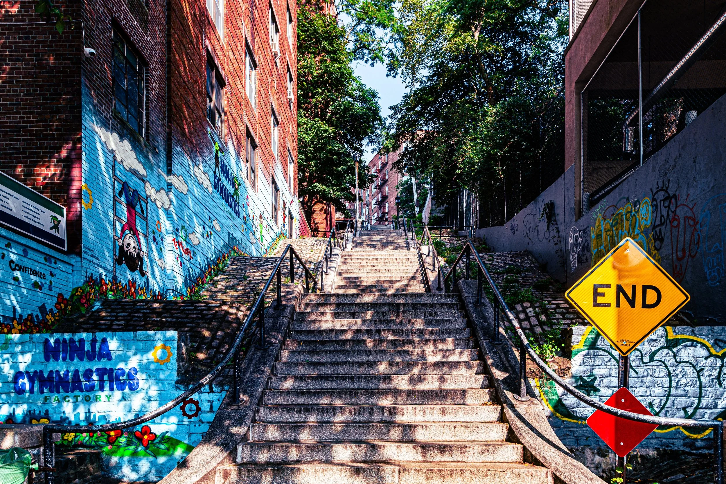 A staircase in an urban area with colorful graffiti murals on the walls on both sides, a yellow 'END' road sign at the bottom right, and trees at the top of the stairs with buildings in the background.