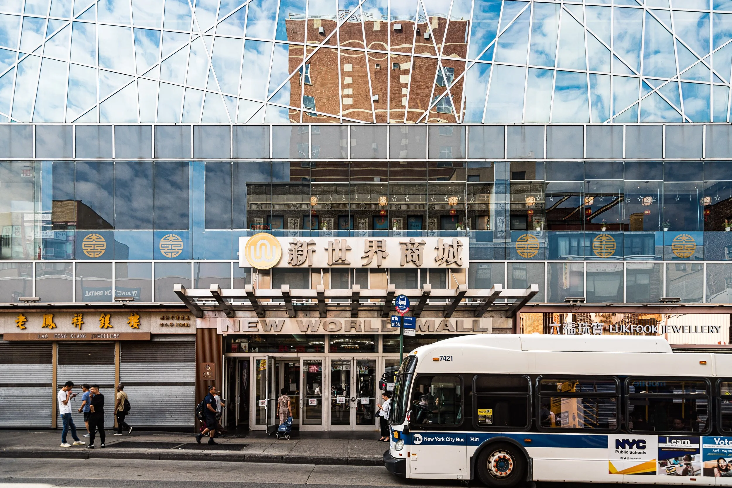 Street scene in front of a shopping mall with people walking and a NYC city bus passing by.