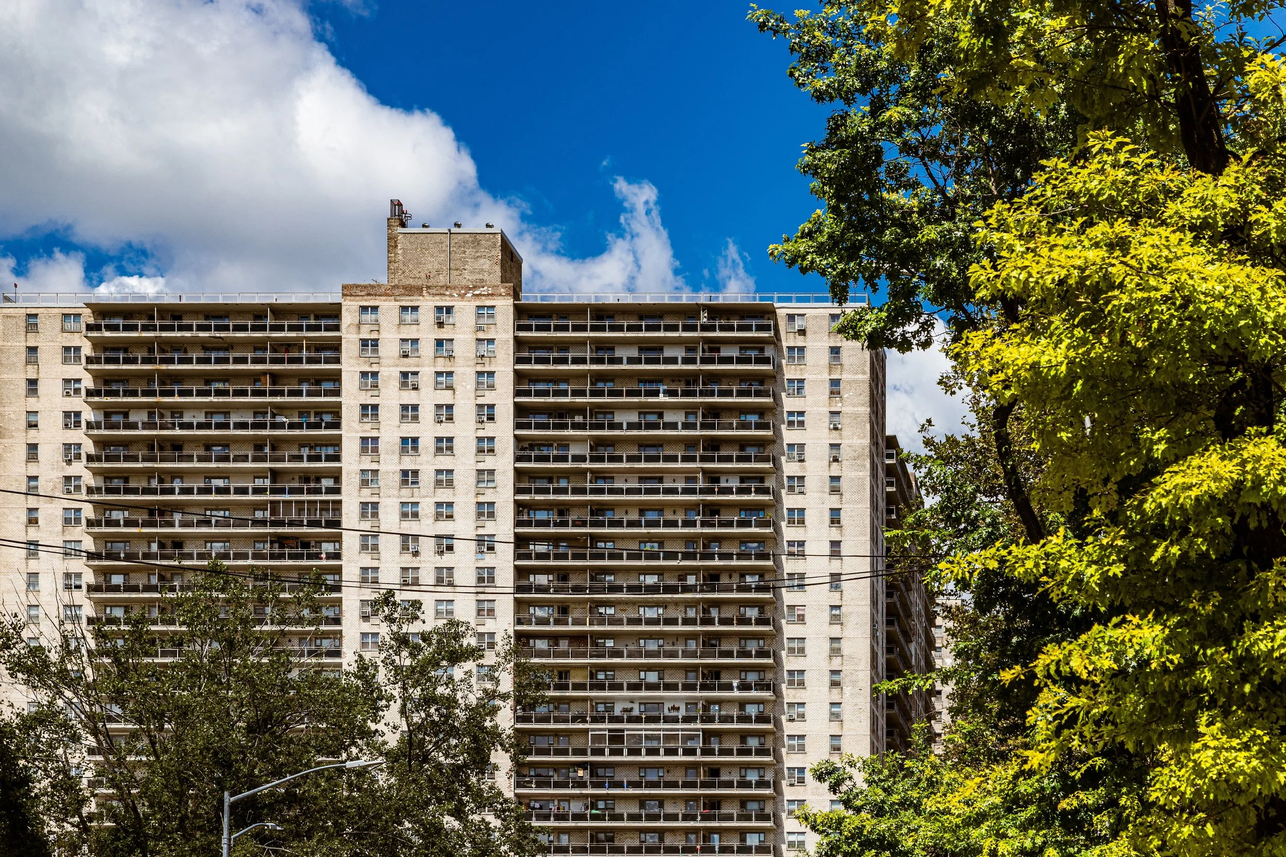 A tall beige apartment building with many balconies, surrounded by trees with green leaves, under a partly cloudy blue sky.