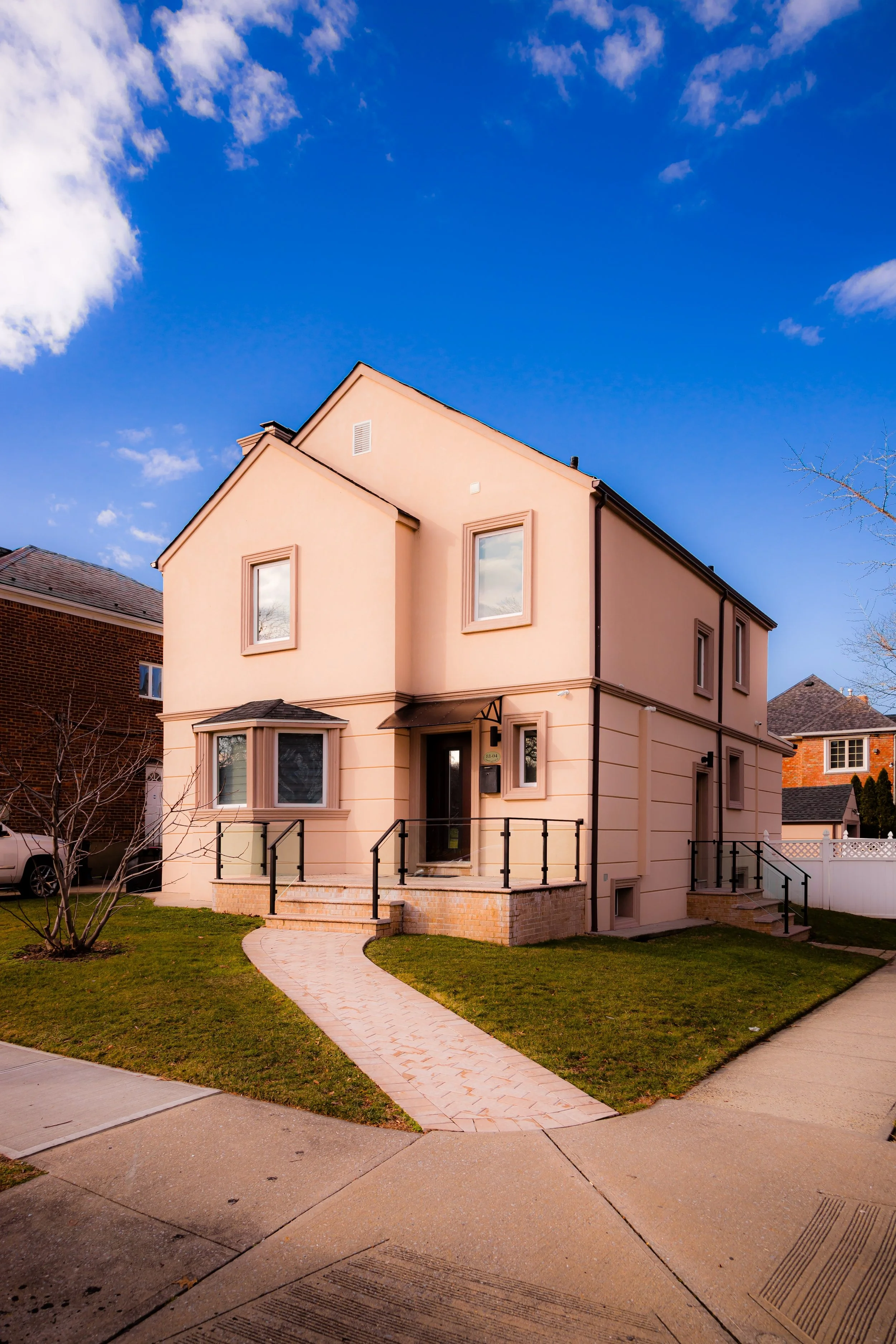 A two-story peach-colored house with front steps, black railings, a brick sidewalk, and a manicured lawn against a partly cloudy blue sky.