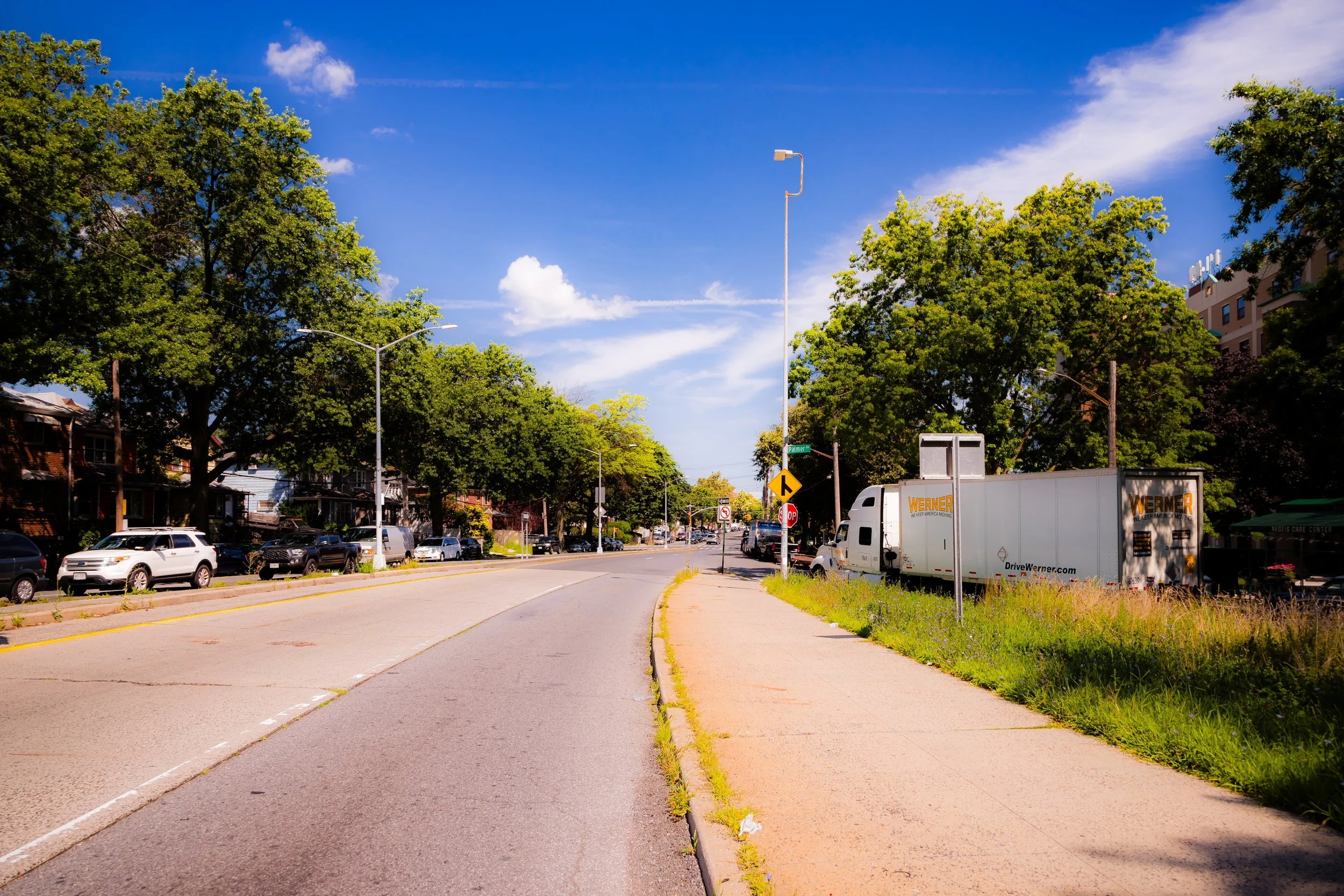 A sunny urban street with parked cars, a delivery truck, trees lining the sidewalks, and a blue sky with some clouds.