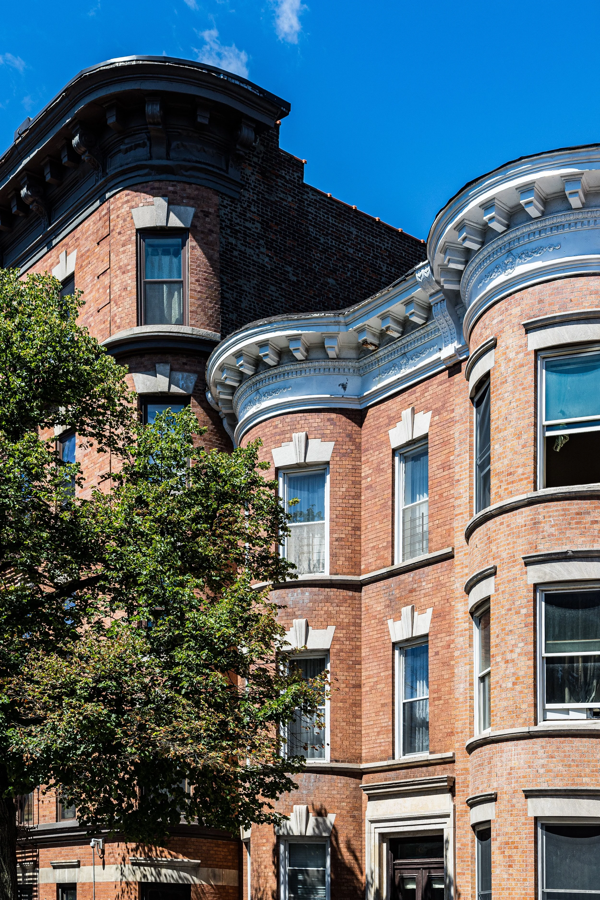 A historic red brick apartment building with ornate white decorative trim, curved windows, and a tree in front under a bright blue sky.