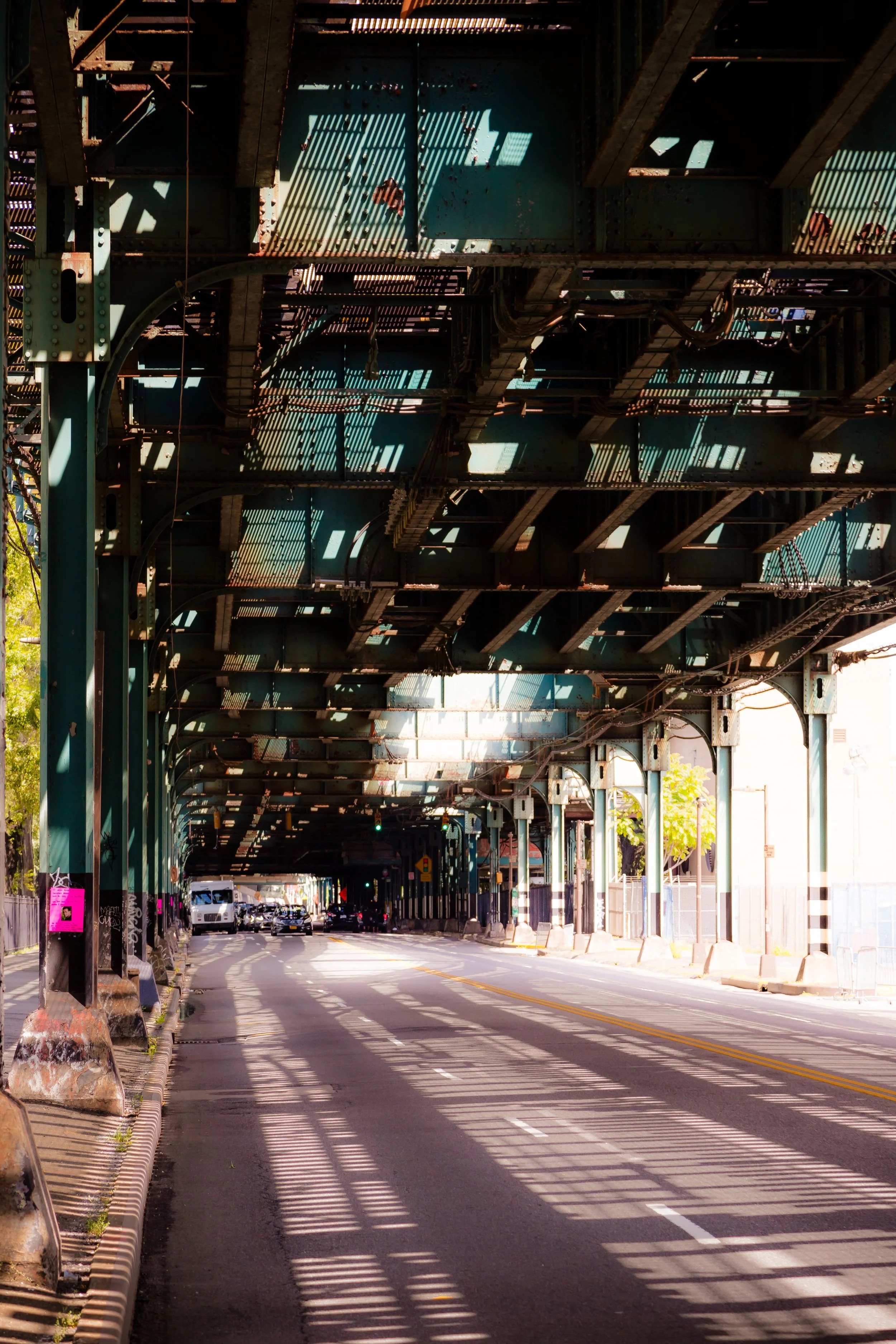 Underneath an elevated train track, a street with cars parked along the curb. Shadows from the train structure create a pattern on the road. Trees and buildings are visible in the background.