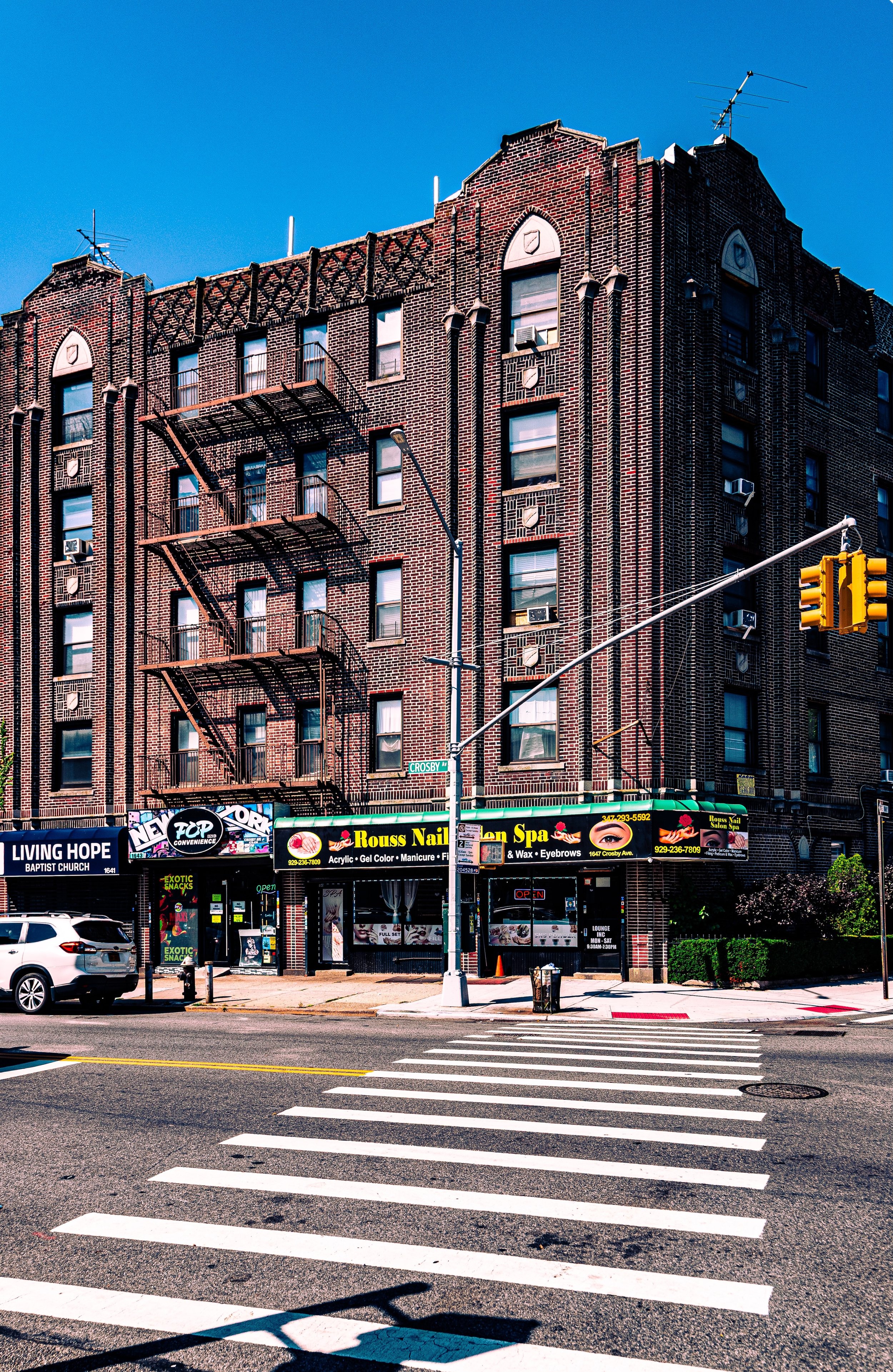 A brick apartment building with multiple fire escapes, retail stores on the ground floor including a nail salon and a convenience store, a pedestrian crosswalk, a traffic light, and a car parked on the street against a clear blue sky.