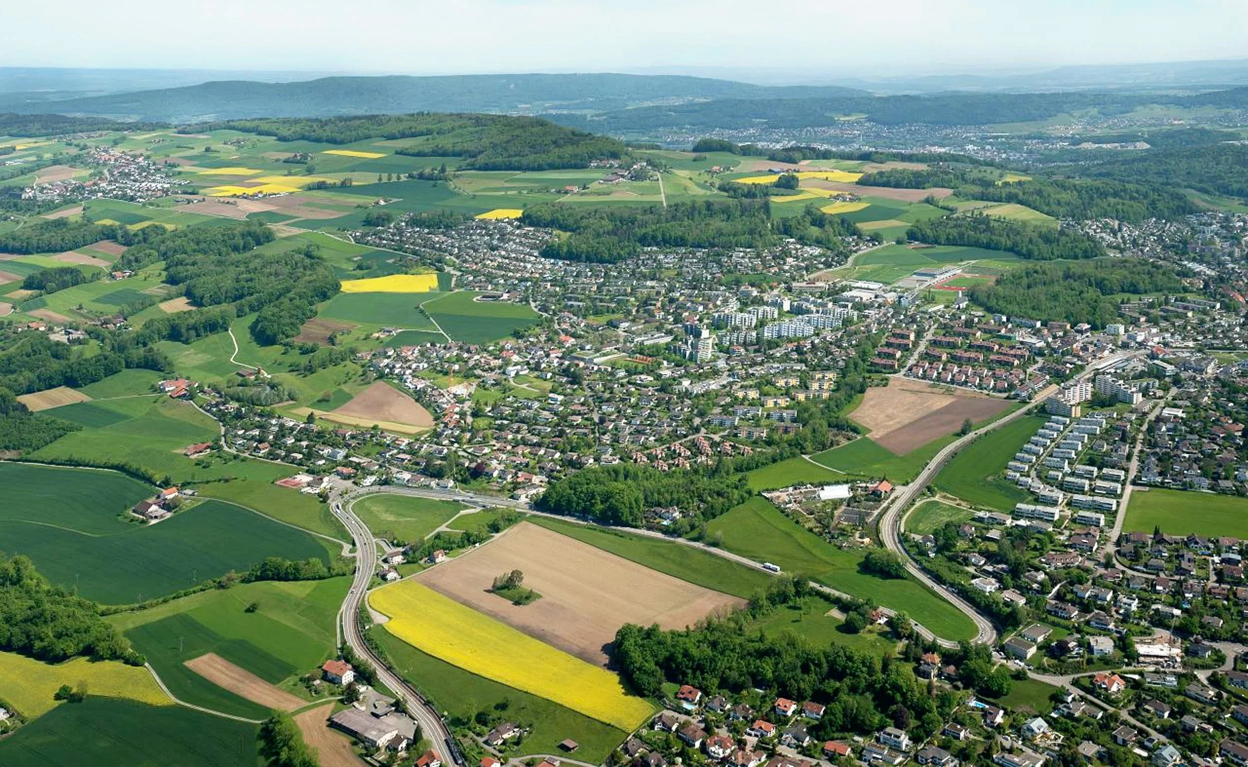 Luftaufnahme einer Stadt mit umliegender grüner Landschaft, Feldern und Straßen.