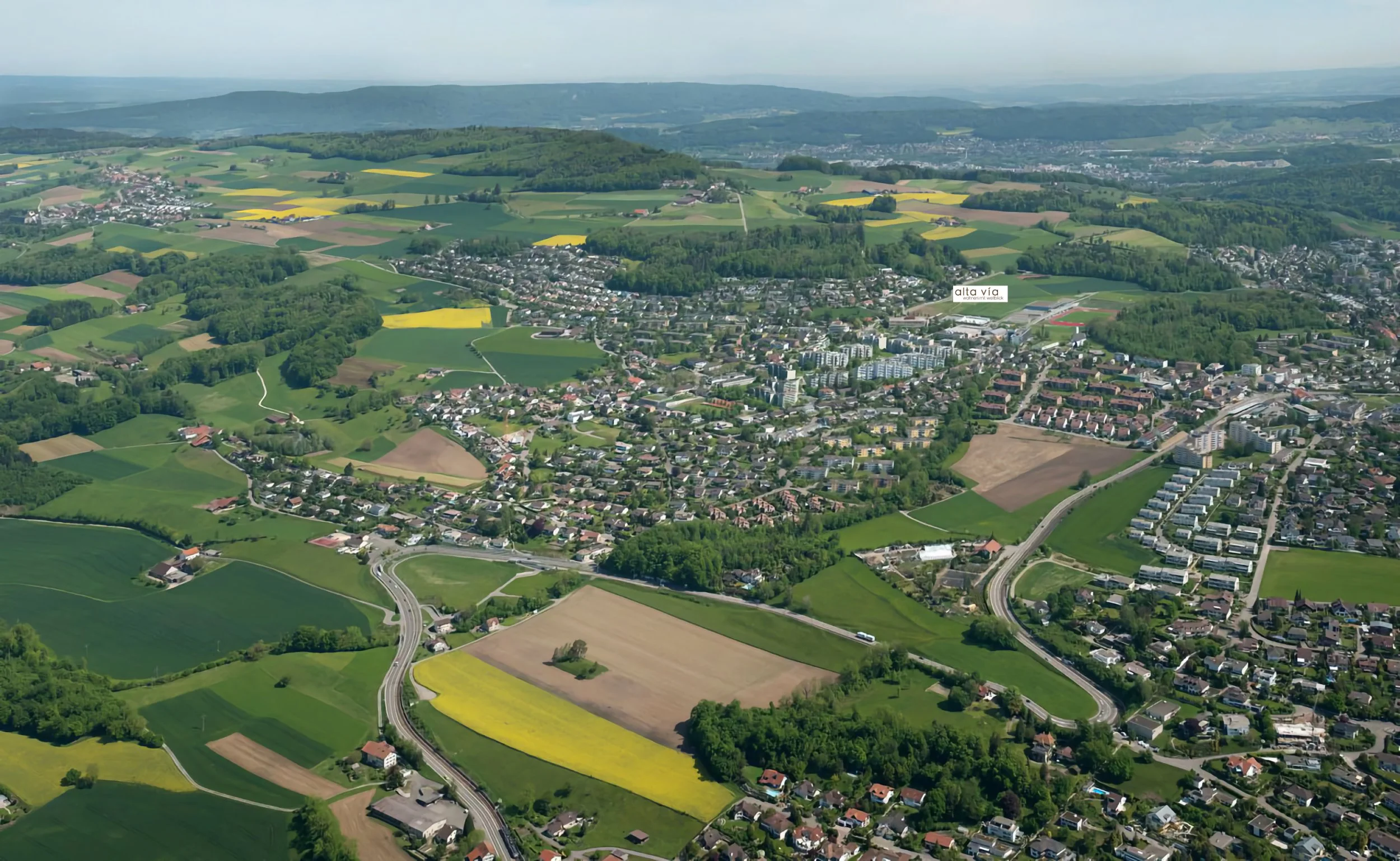 Luftaufnahme eines grünen Hügellands mit einer Kleinstadt, landwirtschaftlichen Feldern, Wäldern und Straßen, mit einem Label 'alta via' im Zentrum. Es handelt sich um eine Luftaufnahmen von Widen AG