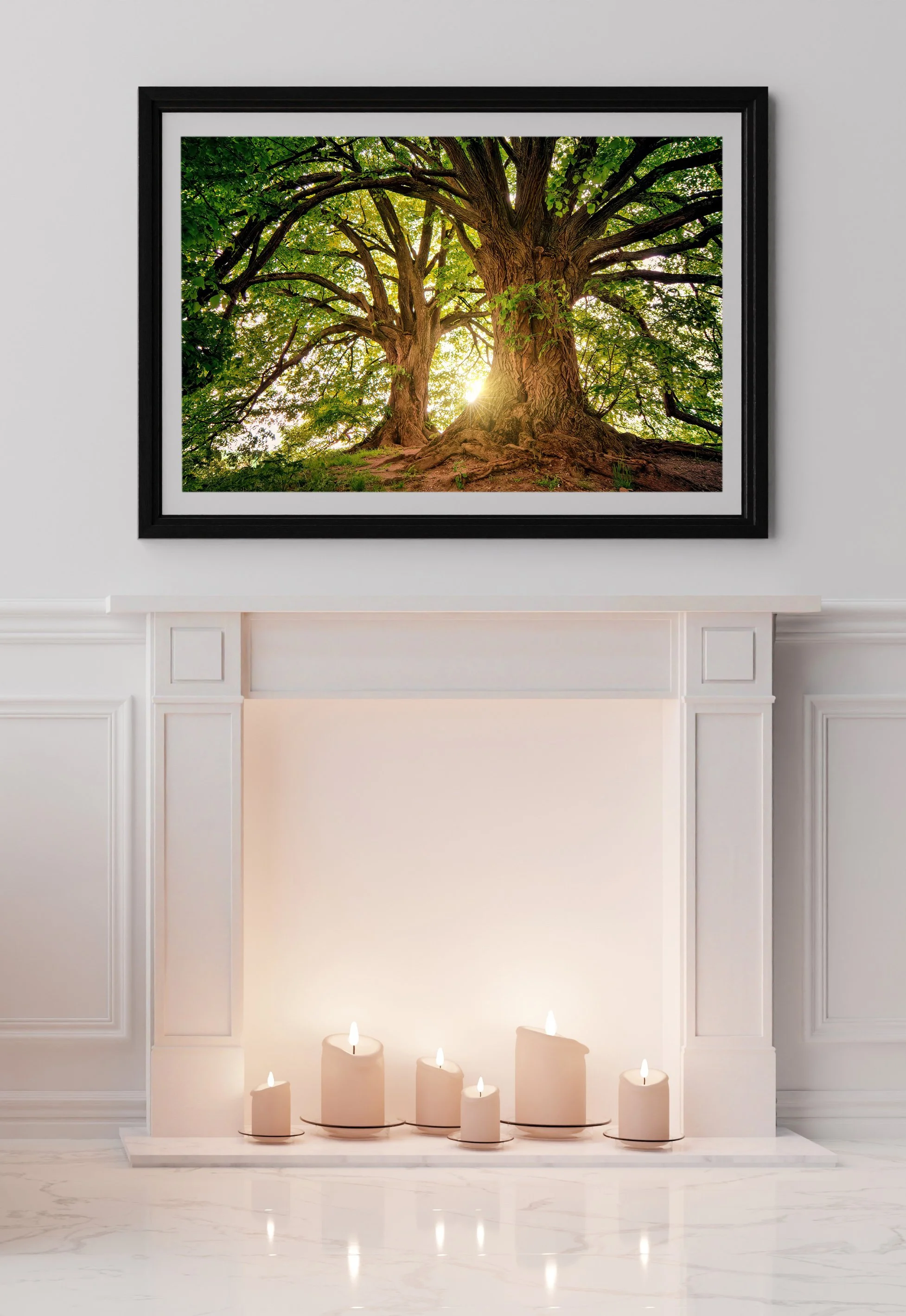 A framed photograph of a large, lush green tree with sunlight shining through the leaves, hanging above a white fireplace with lit candles on the hearth.