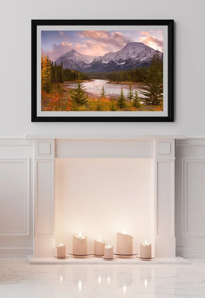 A framed landscape painting of mountains, river, and forest hangs above a white fireplace with candles on the hearth.