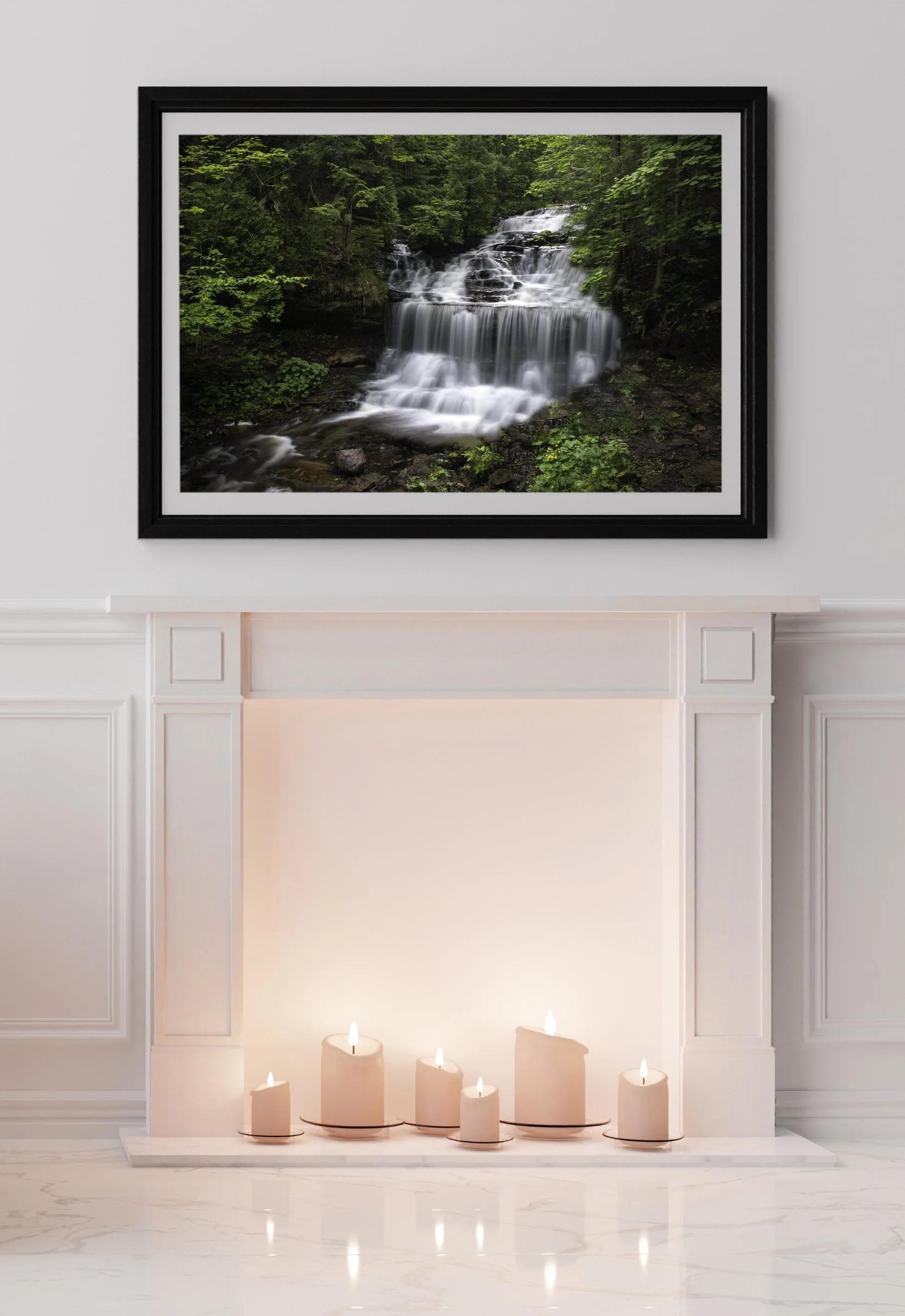 Framed photograph of a waterfall in a forest hangs above a white fireplace with lit cream candles on the hearth.