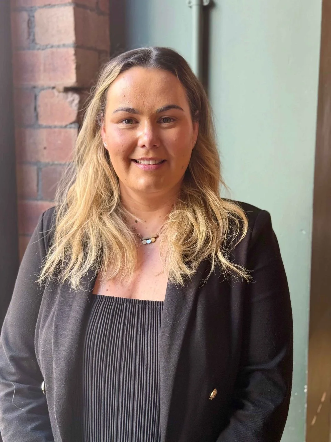 A woman with shoulder-length blonde hair smiling, wearing a black blazer and a black and white striped top, standing indoors near a brick wall and a door.