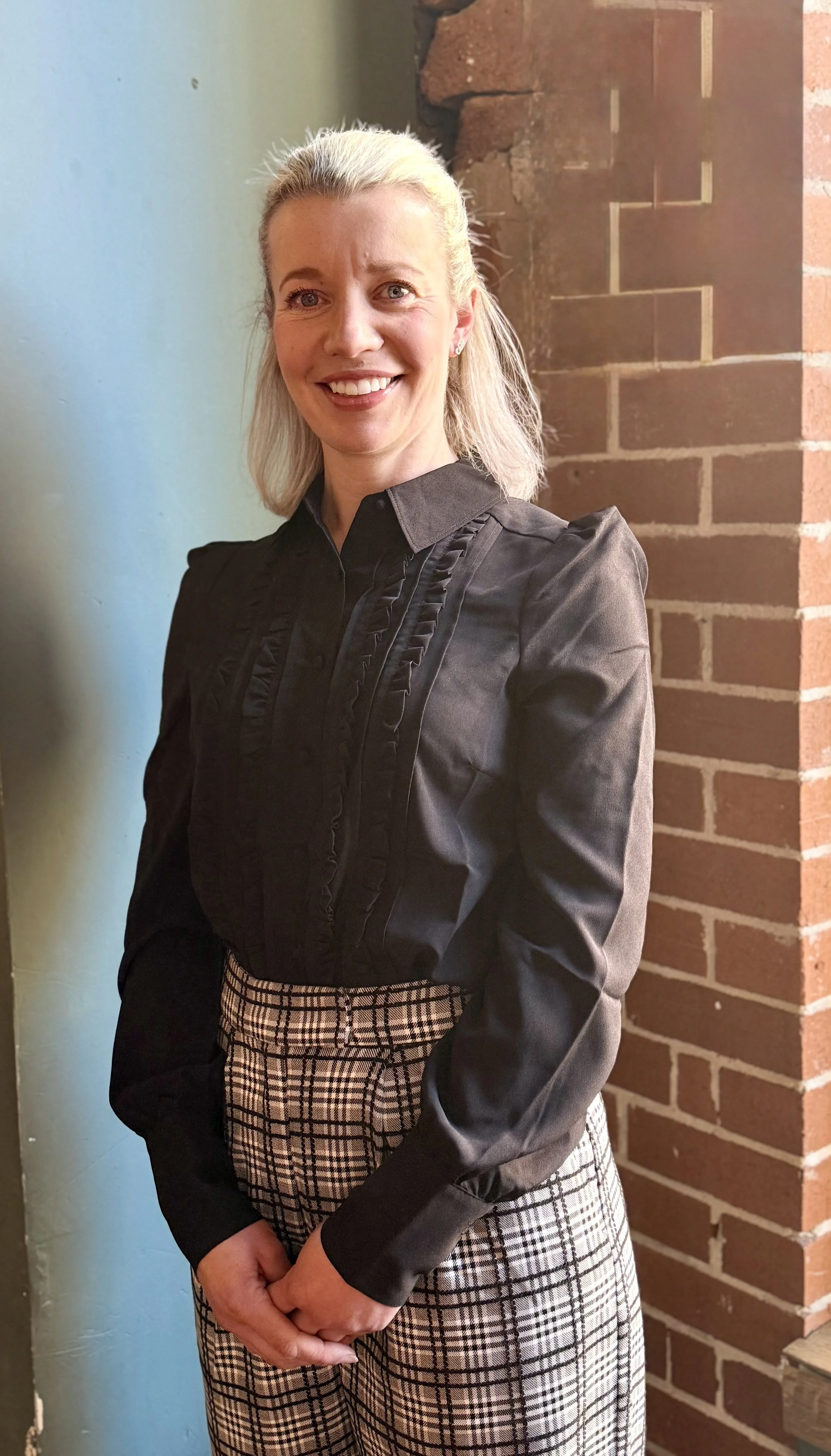Portrait of a woman with blonde hair wearing a black blouse with ruffled details and plaid pants, standing against a brick wall.