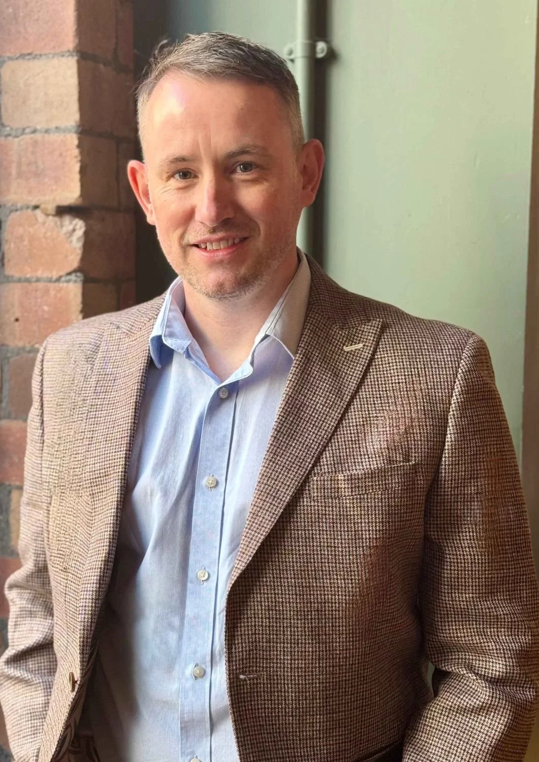 A man with short gray hair and a light beard, smiling, wearing a light blue dress shirt and a brown houndstooth blazer, standing indoors near a brick wall and a green wall.