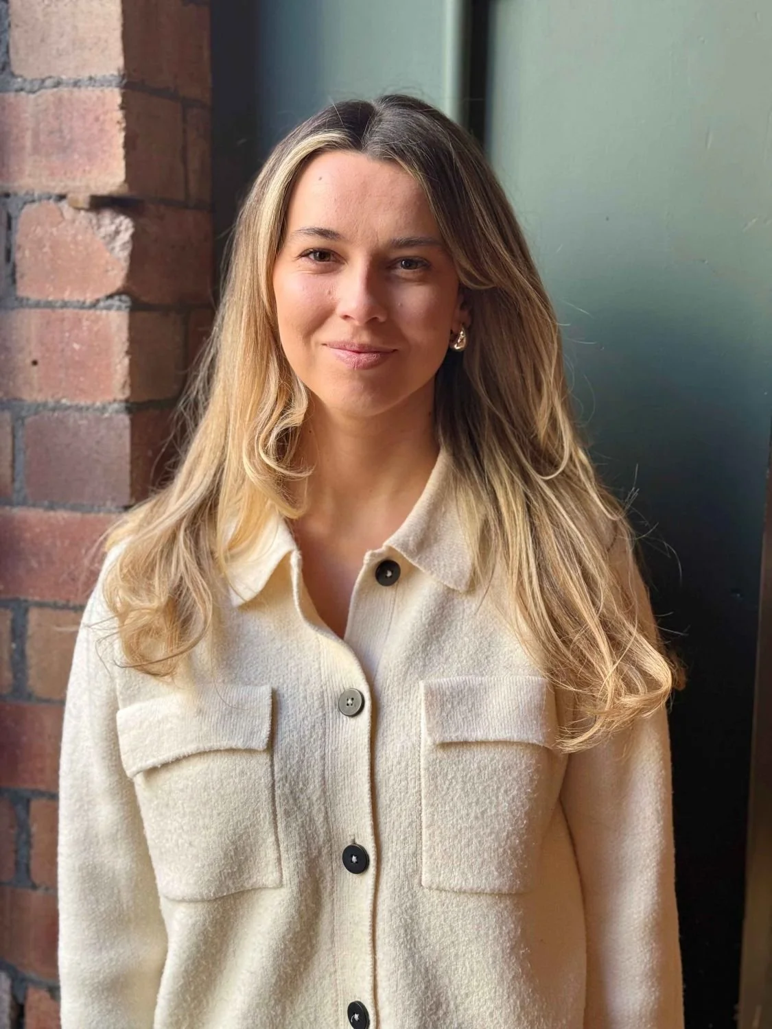 A young woman with long blonde hair standing outdoors near a brick wall and a green door, wearing a beige button-up jacket with two chest pockets, smiling softly.