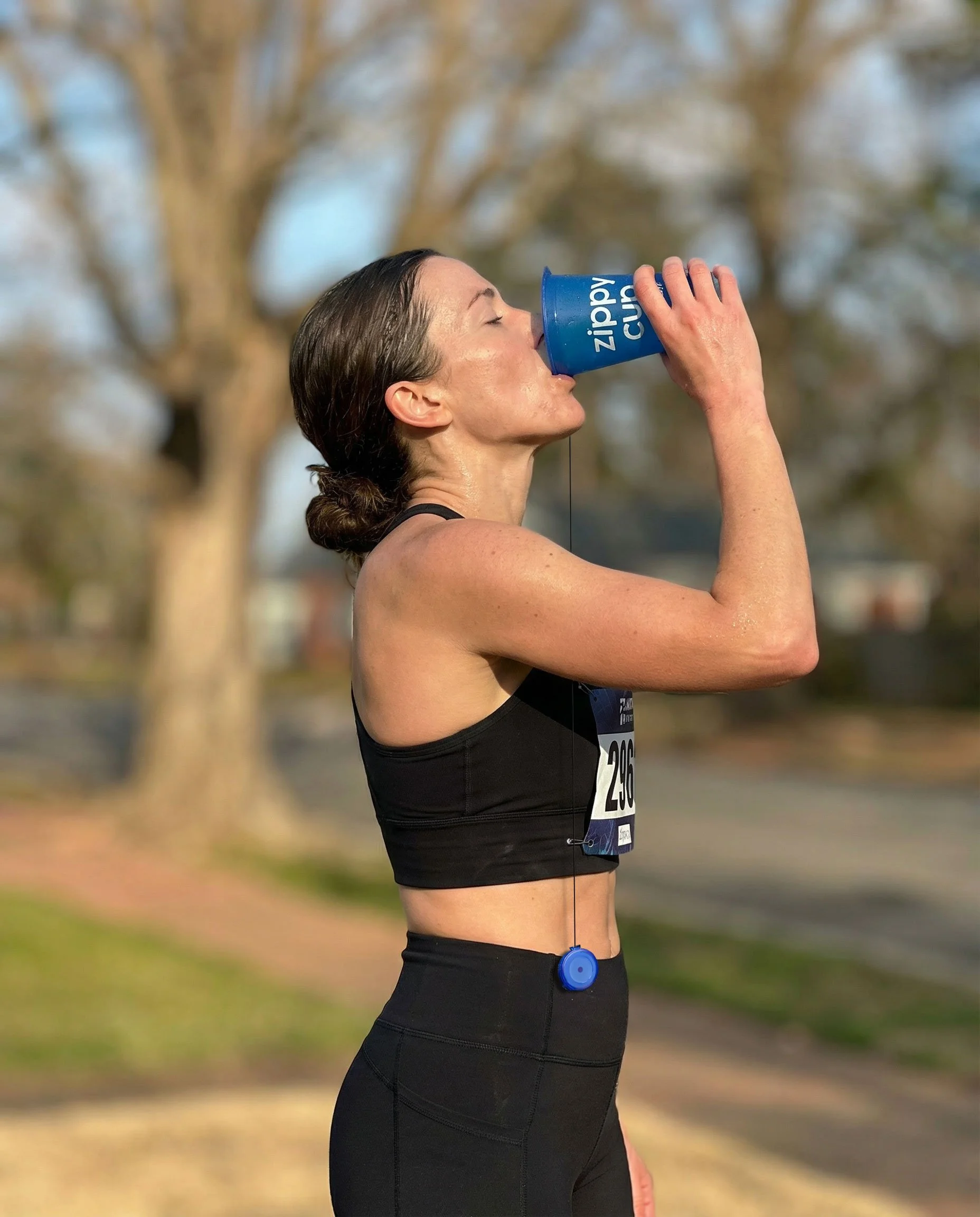 A woman in athletic attire drinking water from a blue zipppy cup during a run outdoors.