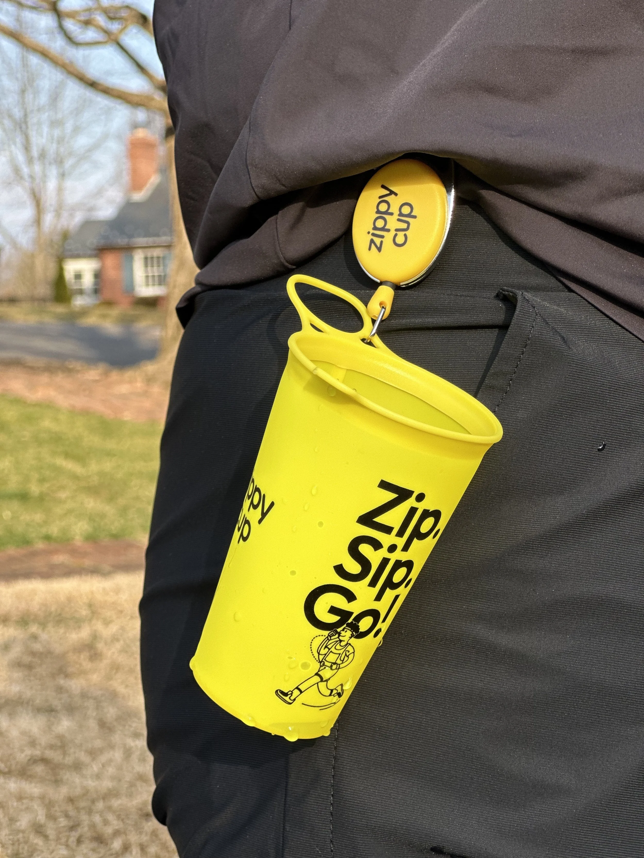 A person wearing dark gray pants and a dark jacket has a yellow Zip S'lip Go cup attached to their waist with a retractable badge reel. The cup has a cartoon character running and the words 'Zip S'lip Go' printed on it.