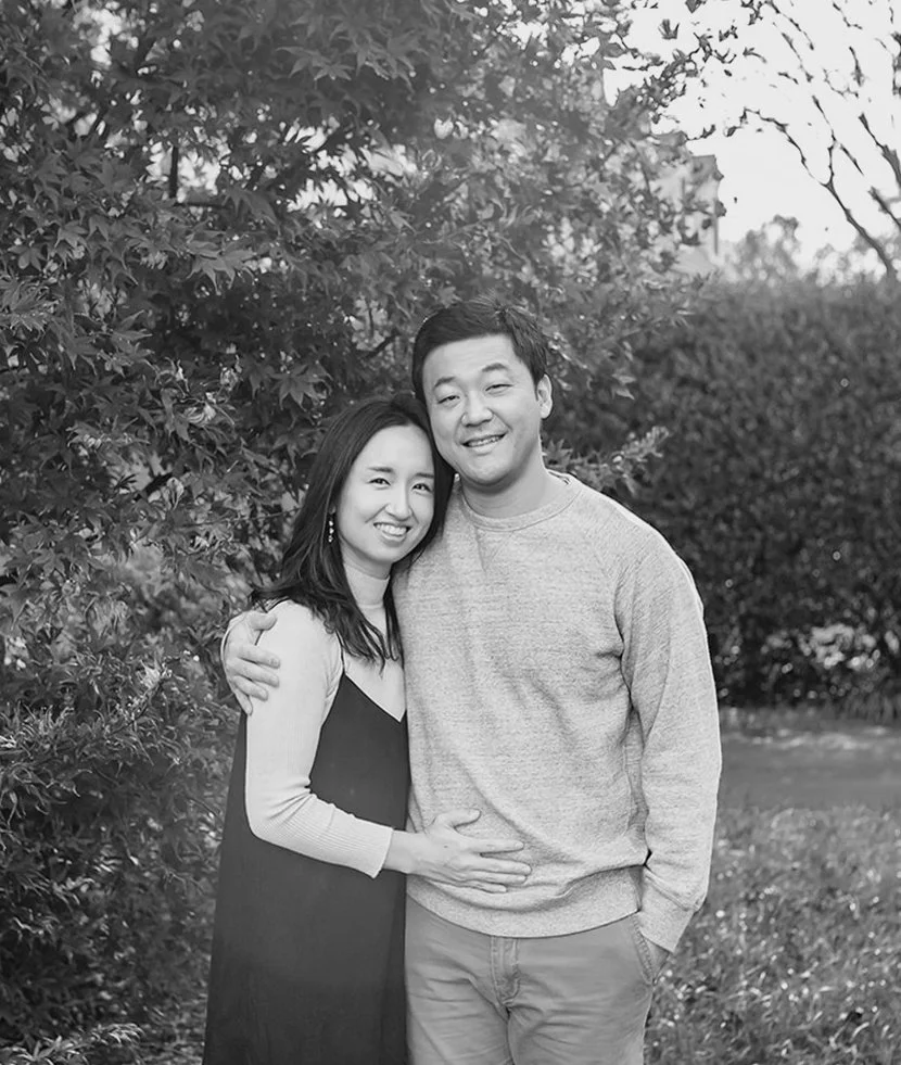 A smiling couple standing outdoors, with the woman hugging the man and the man with his arm around her, in front of trees and bushes.