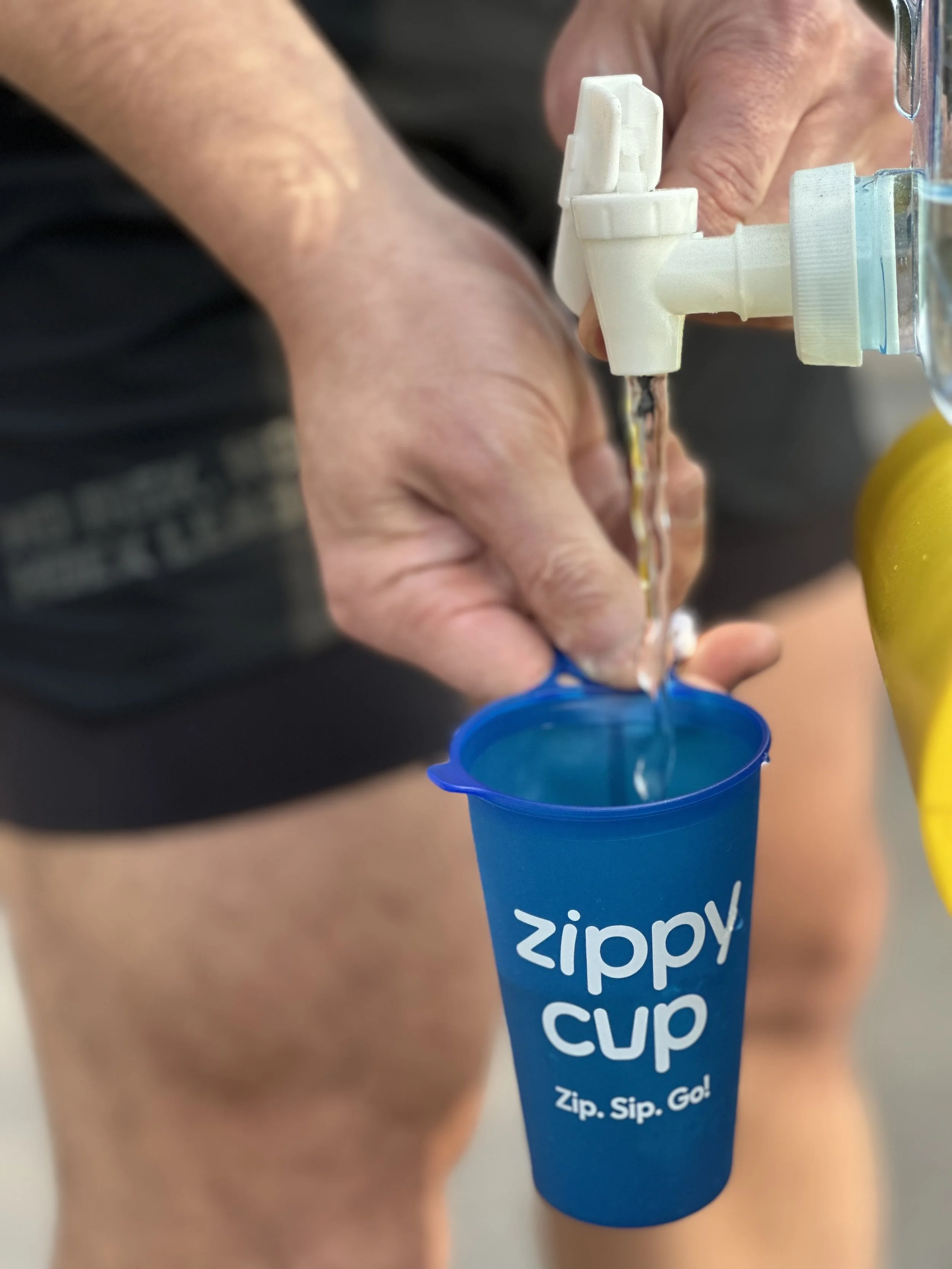 Close-up of a person's hand filling a blue Zippy Cup with water from a water dispenser.
